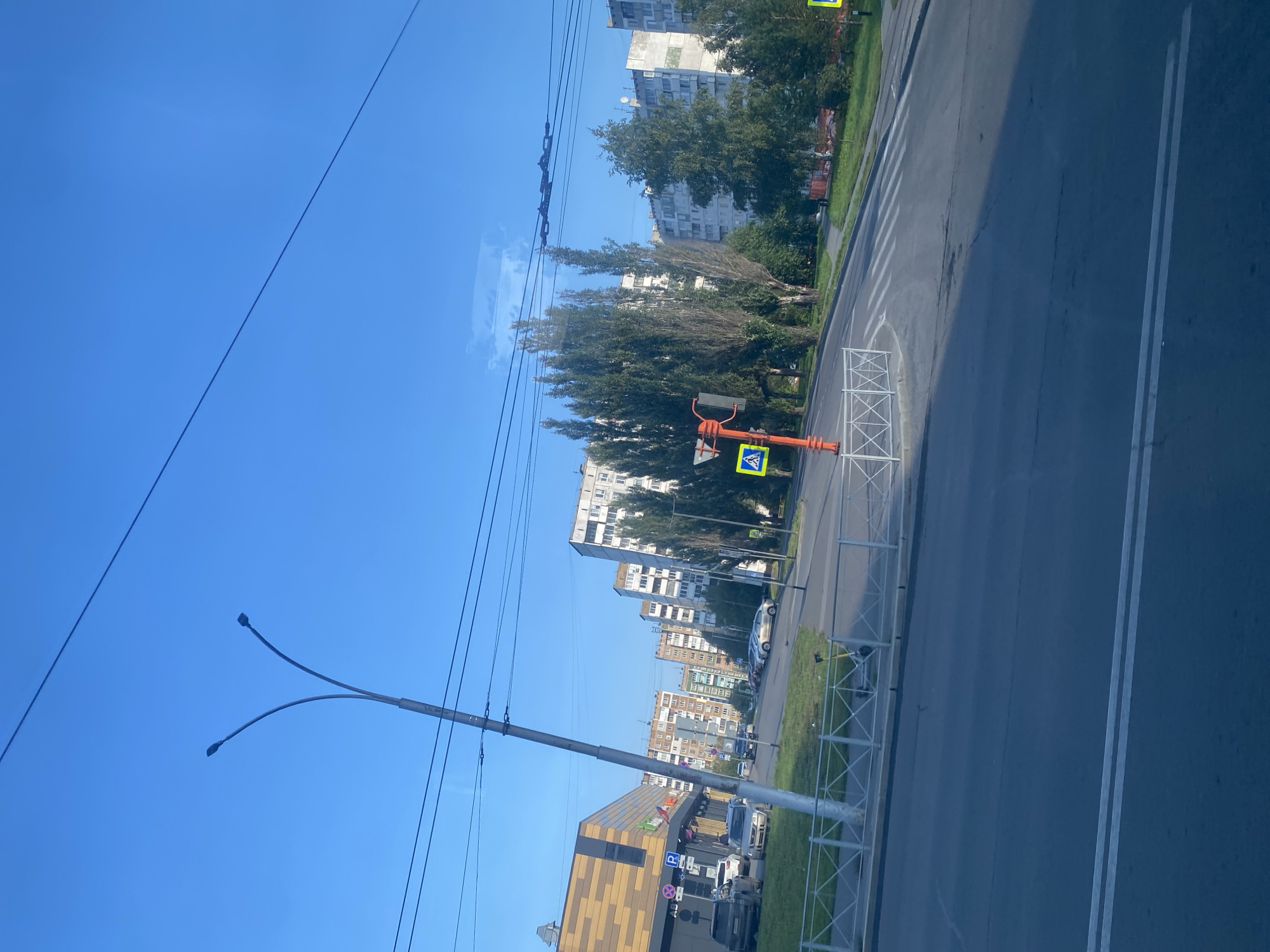 A bright, clear blue sky is visible above a street scene in Kemerovo, Russia. In the foreground, a dark asphalt road with white lane markings stretches into the distance. A pedestrian crossing is marked on the road.
To the left of the road, a modern building with a yellow and brown facade stands next to a parking lot where several cars are parked. A streetlamp with two curved arms, each holding a light fixture, is positioned near the building. A white metal fence runs along the edge of the sidewalk.
Further down the street, a row of tall, multi-story apartment buildings lines the right side of the road. These buildings appear to be of Soviet-era architecture, with balconies and a uniform grey or white exterior. Tall, slender trees with dense green foliage are interspersed between the buildings, casting shadows on the sidewalk.
An orange traffic signal pole with a pedestrian crossing sign is prominently located on the right side of the road, near the pedestrian crossing. Another yellow warning sign is visible further down the road. Overhead, power lines are strung across the sky. The scene appears to be in a residential or mixed-use urban area.
