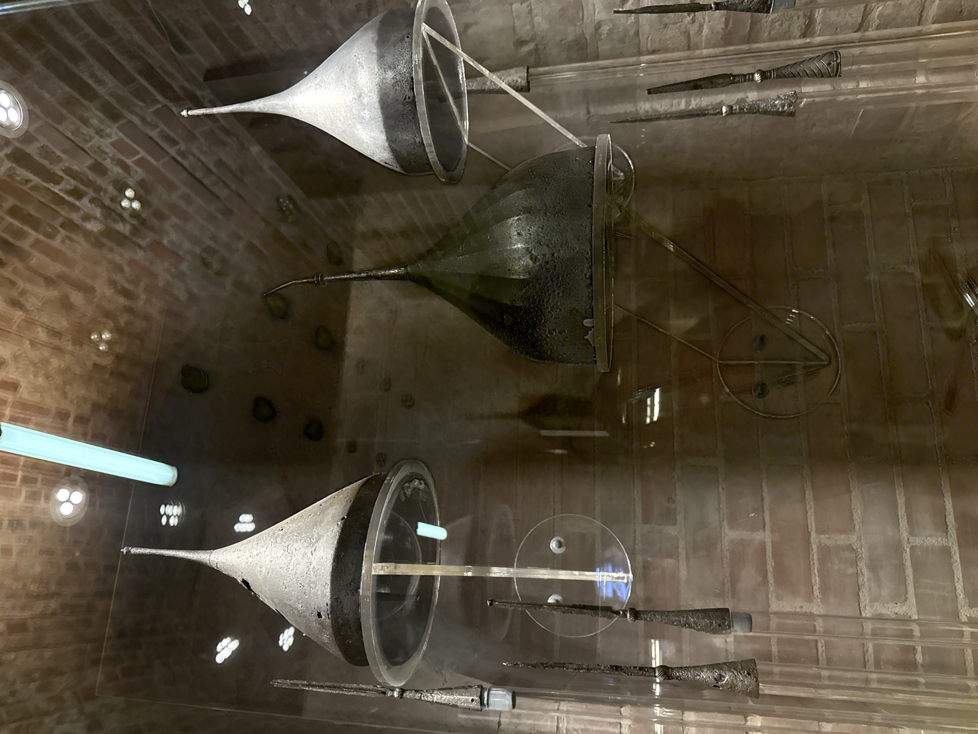 The image depicts items inside a glass display case, likely in a museum or historical building. The background shows a brick wall with embedded light fixtures.
Inside the case, there are several metal objects resembling funnels or cones, along with metallic rods, possibly wands or ornate tools. The arrangement appears to be an exhibit, with some objects seemingly suspended or mounted on transparent stands or rods. The lighting suggests a well-lit interior space. The reflective surfaces within the display make it difficult to discern exact shapes and details, adding a layer of complexity to the scene.
It's not possible to determine the precise location within Moscow based on the image alone. More contextual information would be required to narrow it down.