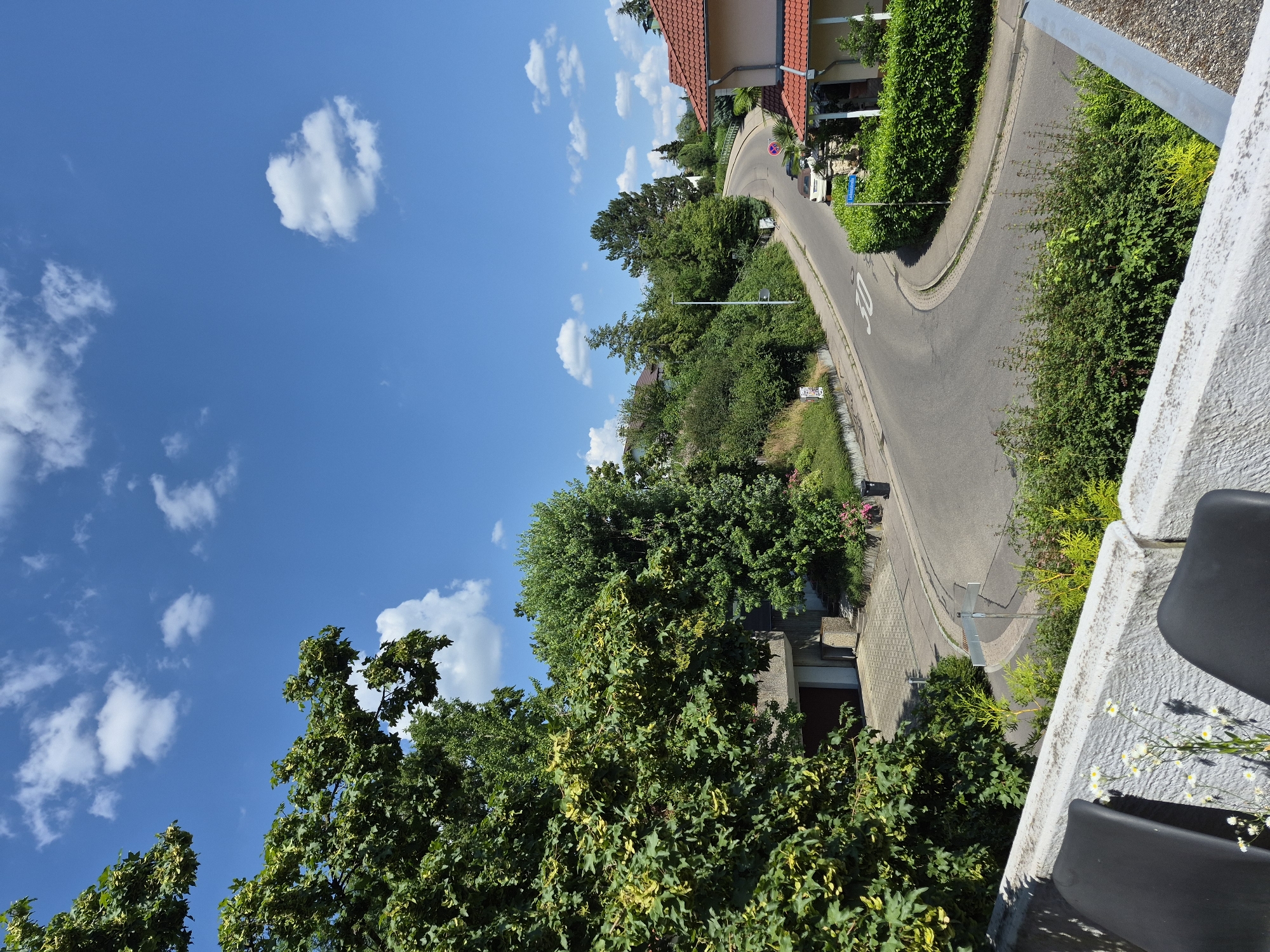 The image depicts a residential street on a sunny day with a clear blue sky and scattered white clouds. A winding asphalt road curves through a neighborhood with houses and lush green trees. In the foreground, a white concrete barrier and two dark chairs are visible, suggesting a balcony or patio overlooking the scene. The houses have red-tiled roofs and are surrounded by well-maintained landscaping, including hedges and flowers. Cars are parked along the street and in driveways. A street sign indicates "Lukasweg." The overall impression is one of a peaceful suburban environment.