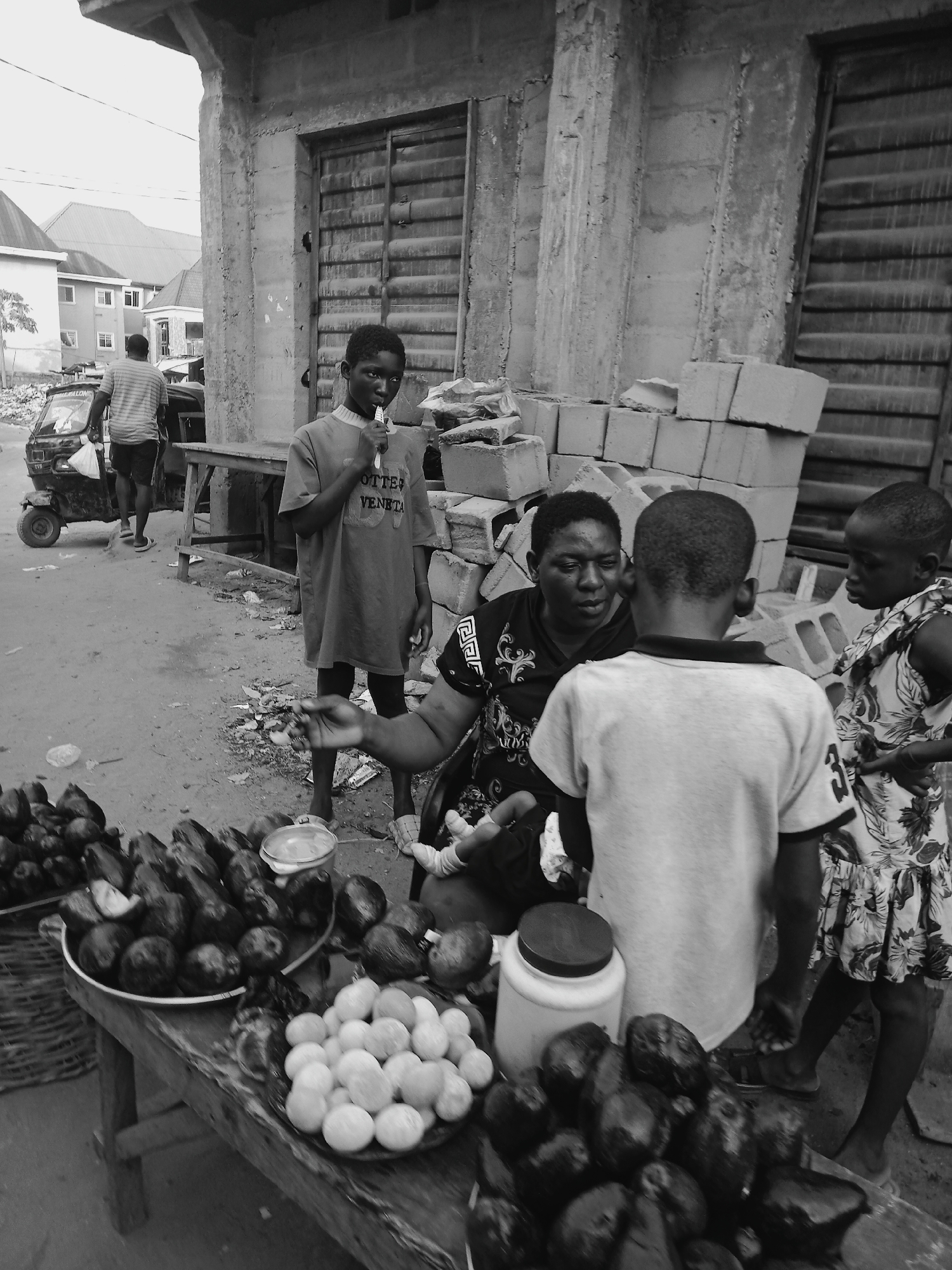 The image depicts an open-air market scene in Onitsha, Nigeria. A woman sits behind a wooden table laden with produce, including avocadoes and what appears to be boiled yams or potatoes. She interacts with two children who stand close to the table. Another child stands nearby, holding what looks like a lollipop. In the background, a man walks past with a small motorized vehicle known as a "Keke Napep." The surrounding area consists of a building under construction, with exposed concrete walls and stacks of cement blocks. The environment suggests a low-income, possibly developing, urban area.