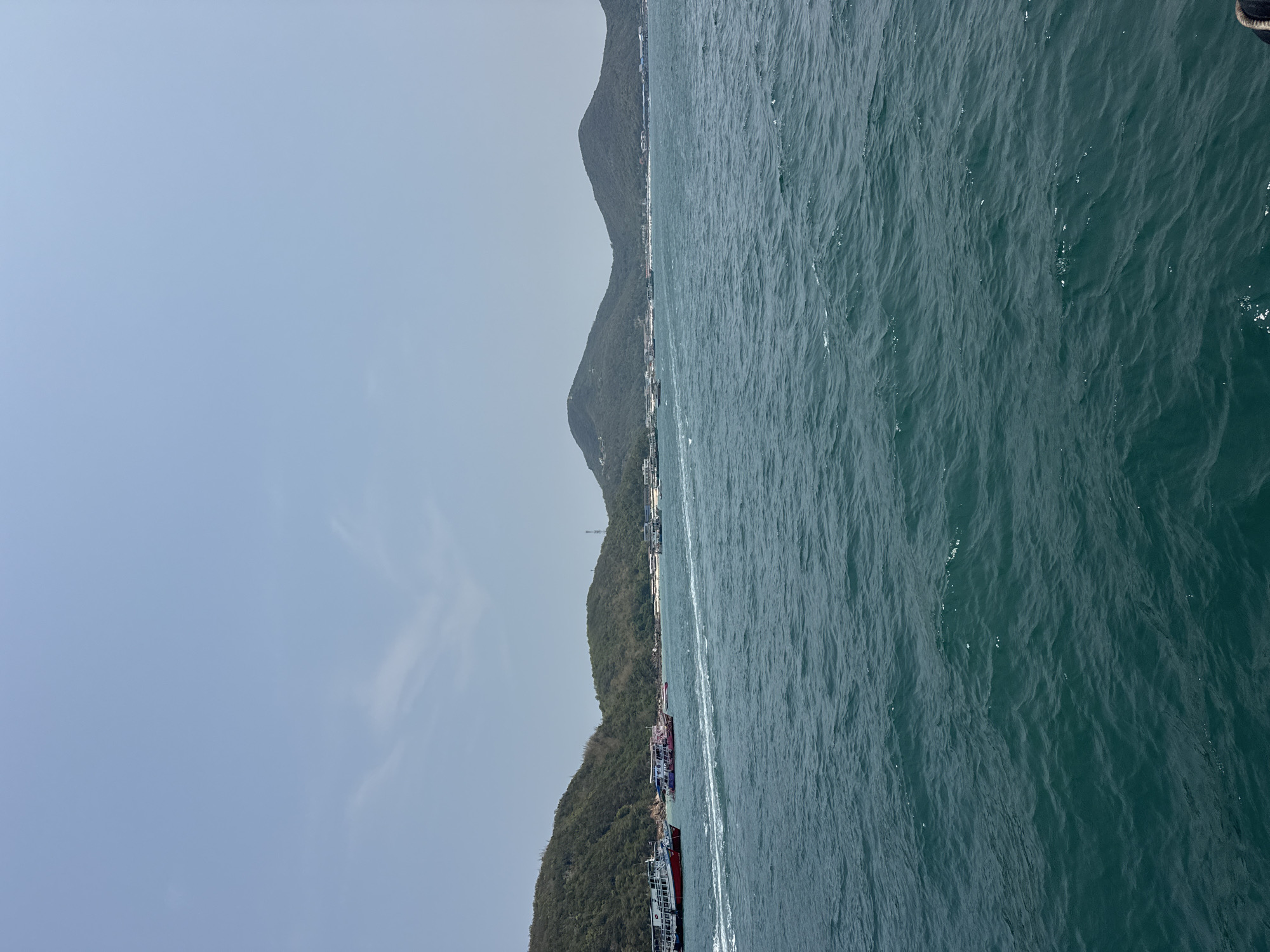 The image captures a view of Pattaya City, Thailand, featuring the ocean, coastline, and hills. The sky is a pale blue with faint cloud formations. The ocean occupies a significant portion of the image and exhibits choppy water with varying shades of blue. The coastline displays a row of buildings and structures. Further inland, three hills are densely covered in green vegetation.
