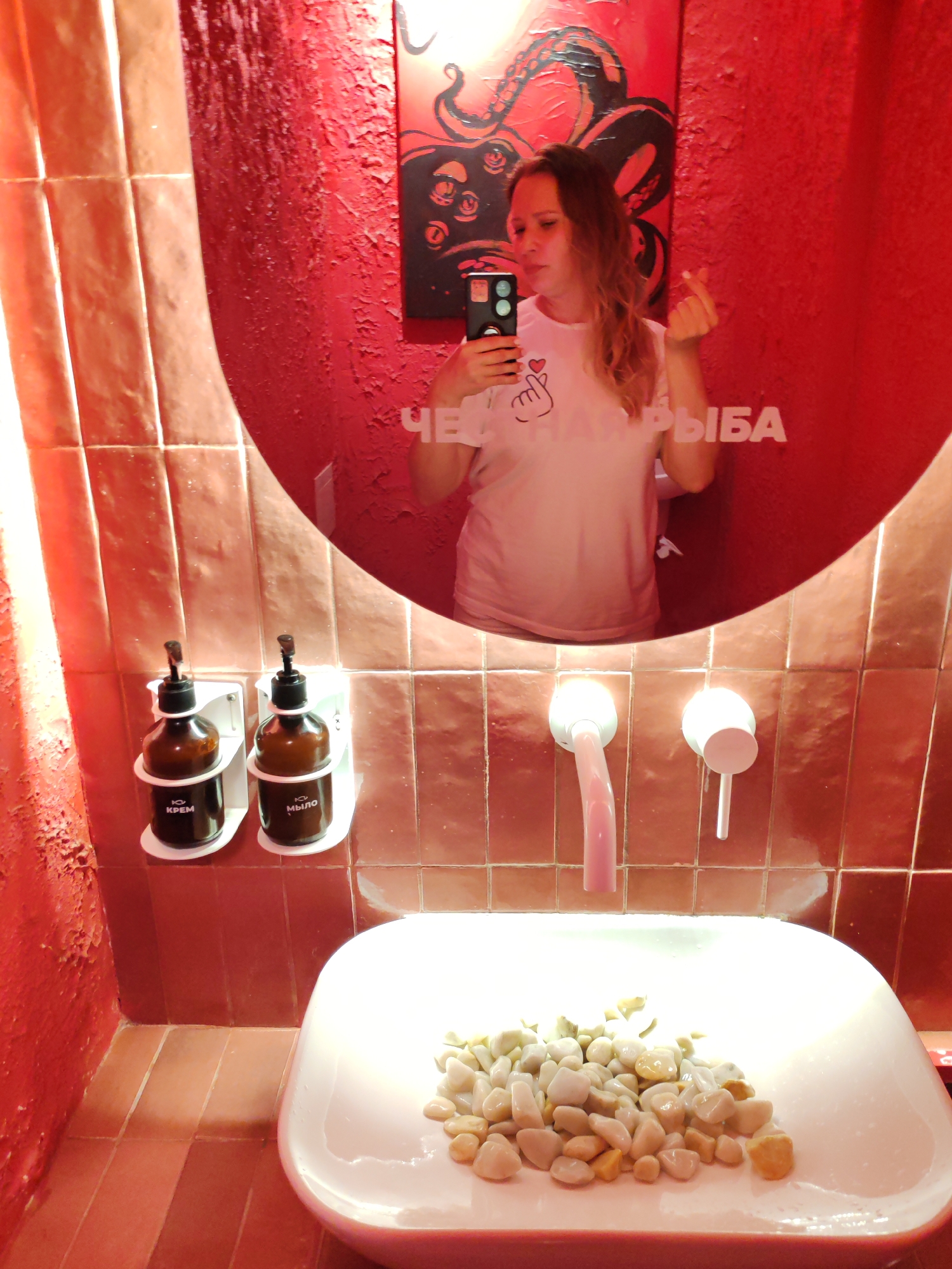 This is a vertical, medium shot photograph taken in a brightly lit restroom with predominantly red decor. A young woman with blonde, wavy hair is reflected in a large, oval mirror. She is holding a smartphone in her right hand, taking a selfie, and making a heart gesture with her left hand. She is wearing a white t-shirt with a pink graphic of hands forming a heart. Her expression appears playful.
The restroom has reddish-brown tiled walls and a red textured wall visible behind the woman in the mirror. Below the mirror, mounted on the tiled wall, are two brown pump bottles, one labeled "КРЕМ" (cream) and the other "МЫЛО" (soap). To the right of the bottles are two modern, white faucets.
In the foreground, a white sink is partially visible, filled with an assortment of smooth, light-colored pebbles.
The overall atmosphere is modern and quirky, with the bold red color scheme and the decorative pebbles in the sink adding unique touches. The image appears to be taken indoors, likely during the daytime, given the strong lighting. Text visible in the reflection of the mirror spells out "ЧЕ" followed by "РЫБА" which translates to "WHAT FISH".