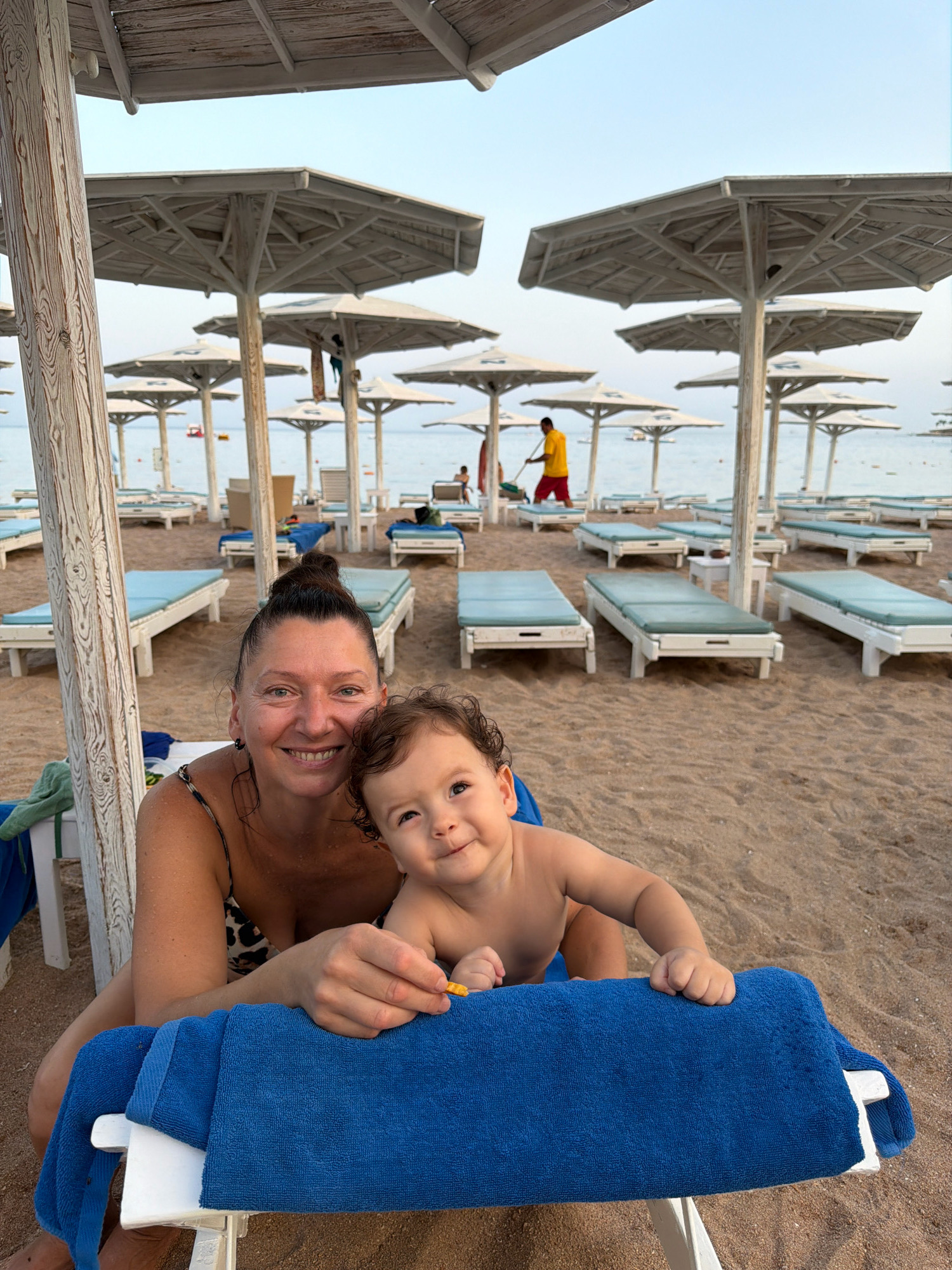 A woman and a baby are sitting on beach chairs under a beach umbrella at a resort in Sharm El Sheikh, Egypt. The woman is smiling and looking at the camera. The baby is looking at the camera and smiling. The beach is made of sand. There are lounge chairs and beach umbrellas on the beach. The ocean is in the background. The sky is clear and blue.