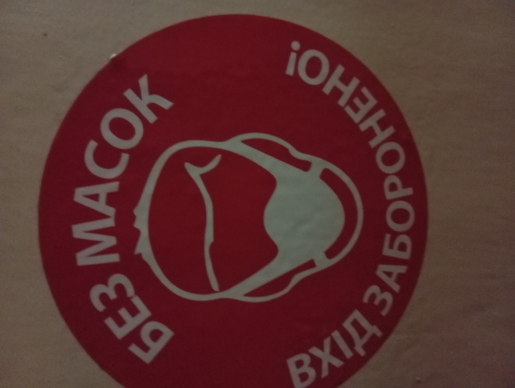 The image is a close-up, slightly angled shot of a red circular sticker with white text and graphics. The sticker depicts a simplified outline of a person's head wearing a face mask. The text on the sticker is in Ukrainian. The top arc of the circle reads "БЕЗ МАСОК" (Bez masok), which translates to "Without masks." The bottom arc of the circle reads "ВХІД ЗАБОРОНЕНО!" (Vkhid zaboroneno!), meaning "Entry is forbidden!" The sticker is affixed to a light-colored, textured surface, possibly cardboard or a wall. The lighting is dim and appears to be artificial, creating shadows and a slightly grainy texture. There is no specific information in the image to determine its exact location within Zaporizhzhia, Ukraine. The sticker is a clear visual warning indicating that entry is not permitted for individuals not wearing face masks.