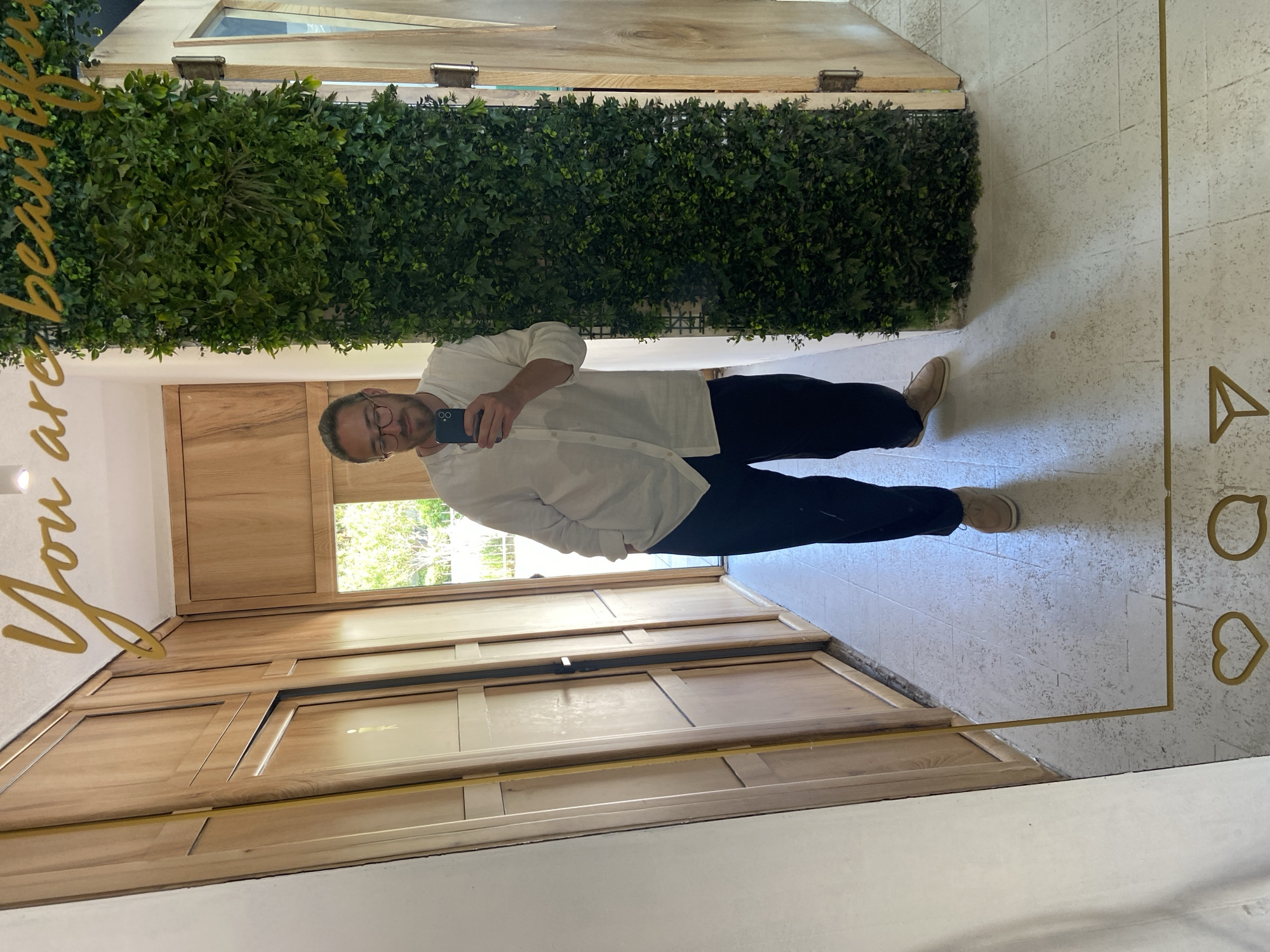 A middle-aged man takes a picture of himself in a large mirror. The mirror is framed with gold markings resembling social media icons and the words "You are beautiful." The man wears glasses, a white button-down shirt, and black pants. The background consists of wooden cabinets, a patch of greenery, and what appears to be an opening to the outdoors, suggesting a garden or courtyard. The location is Petrovac centar, Montenegro.