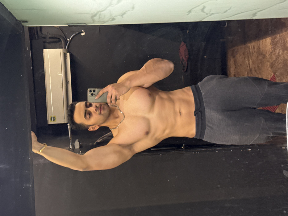 A muscular man poses for a mirror selfie in what appears to be a gym or fitness setting. He's shirtless, revealing defined chest and abdominal muscles, and wears dark grey sweatpants. He holds a smartphone in his right hand, capturing the image. A gold bracelet is visible on his left wrist, and a silver chain around his neck. The background features dark walls with possible gym equipment or fixtures partially visible. The overall image emphasizes the man's physique and fitness level.