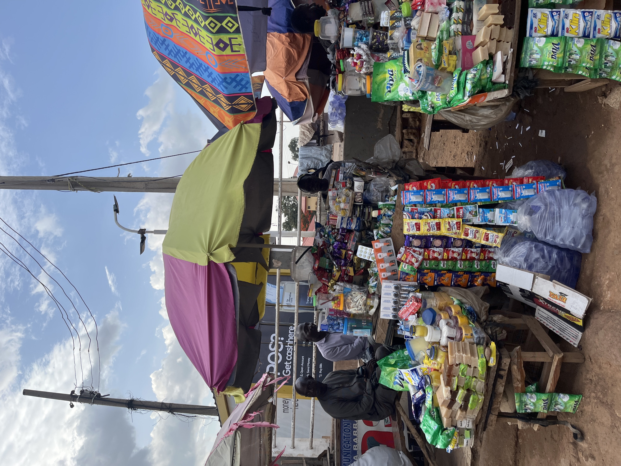 The image depicts an outdoor market stall in Gura Top, Nigeria. The stall is covered by colorful canopies providing shade. Two men are sitting behind the stall, possibly vendors or customers. The stall is stocked with various goods, including soap, detergent, packaged snacks, and bottled water. A sign on the stall reads "POS Get cashhere". There are power lines visible above the stall. The scene is bustling with activity, suggesting a busy marketplace.