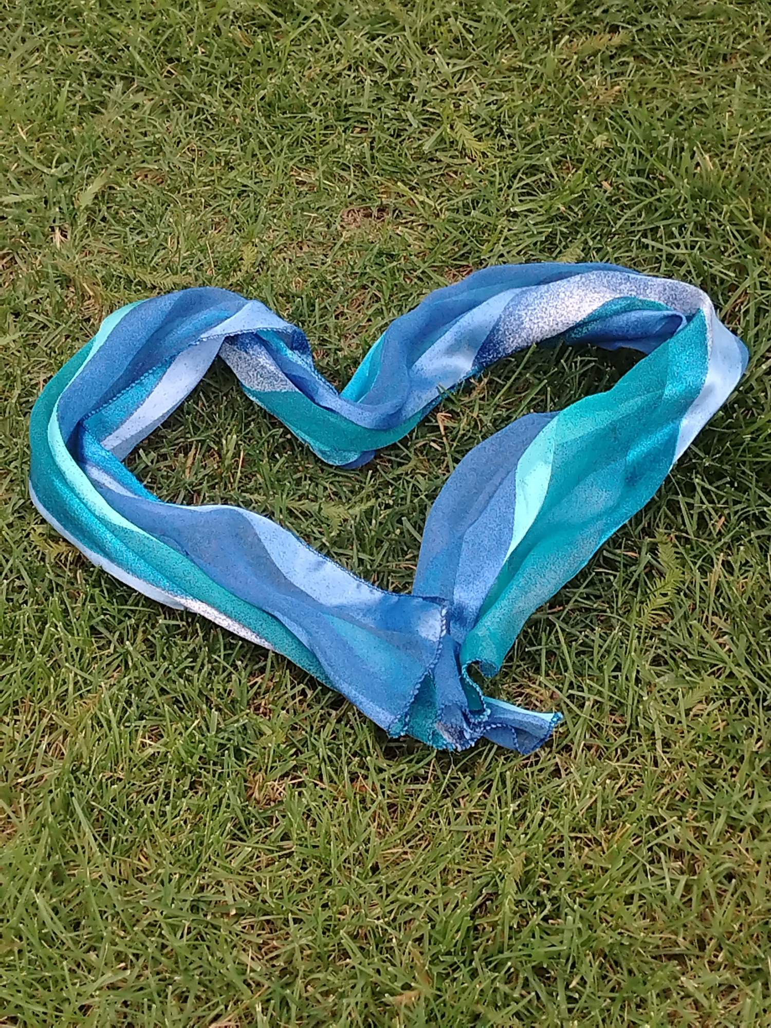 A light blue and turquoise scarf with wave-like patterns and some speckled detailing is arranged in the shape of a heart on a bed of green grass. The scarf is made of a lightweight, flowing material, possibly silk or chiffon. The grass appears somewhat dry in places, with some patches of yellowing. The image is taken from a top-down perspective. There are no people or any other objects visible in the frame. The scene is outdoors, with natural lighting. The location is not specified as being in the city of Krasnodar, Russia.
