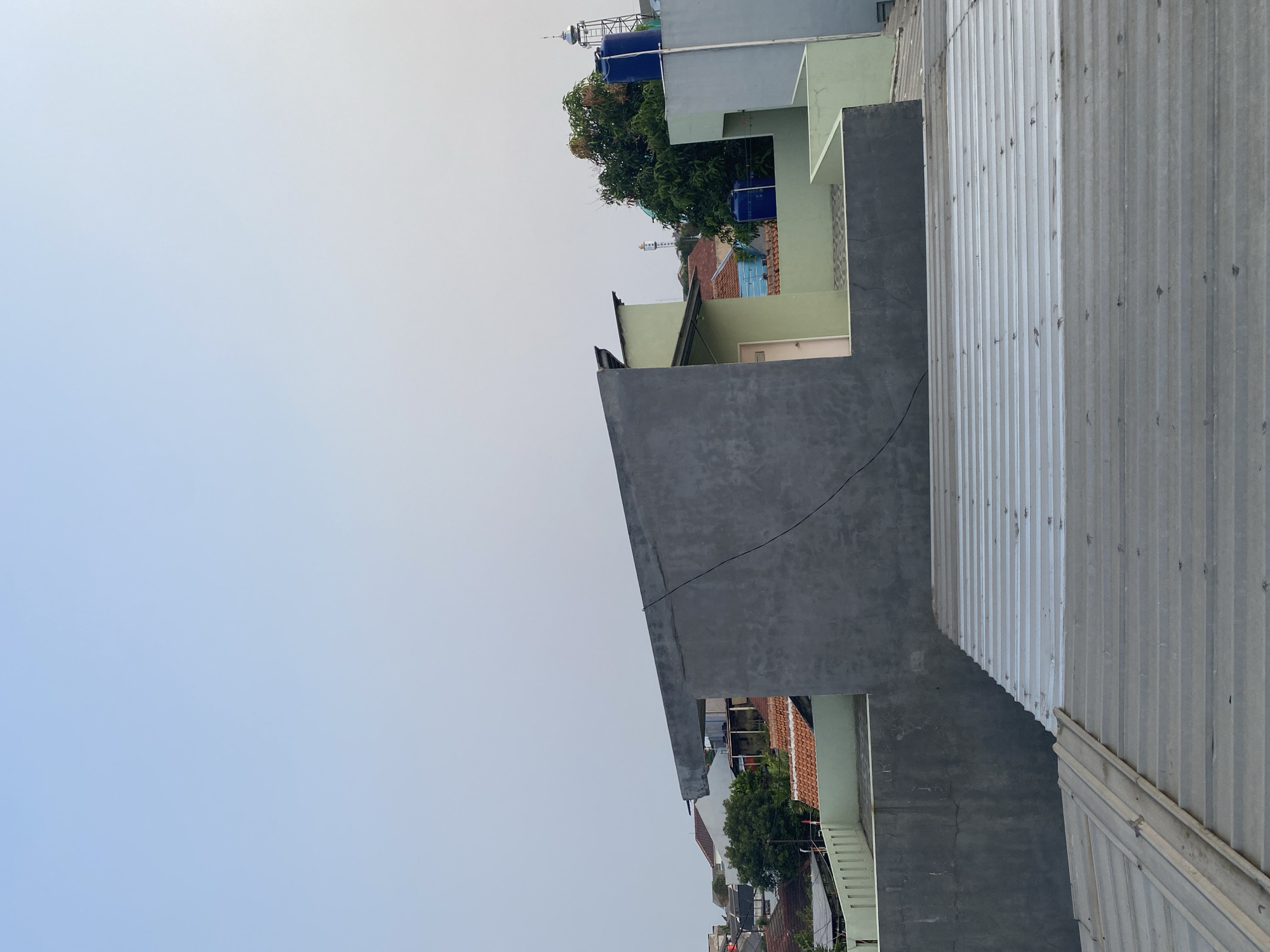 The image captures a view of rooftops in a densely populated urban area, likely a residential neighborhood in Tangerang, Indonesia. The foreground features corrugated metal roofs, one of which is partially covered by a lighter-colored corrugated material. Behind these, a grey concrete wall extends upwards, topped with a concrete overhang.
Beyond this structure, a cluster of buildings is visible. A light green building with a white door is prominent, with its side wall forming a corner. Adjacent to it, another building is painted a pale blue, and further to the right, a light grey building with a light blue water tank on top is partially visible. Red-tiled roofs are interspersed among the buildings, characteristic of Indonesian architecture.
Green trees are scattered throughout the scene, adding a touch of nature amidst the concrete and metal structures. In the background, a tall tower, possibly a communication tower or a water tower, rises above the rooftops, with a mosque minaret also discernible. The sky is a pale, hazy blue, suggesting either early morning or late afternoon light. The overall impression is of a typical urban landscape with modest residential buildings.