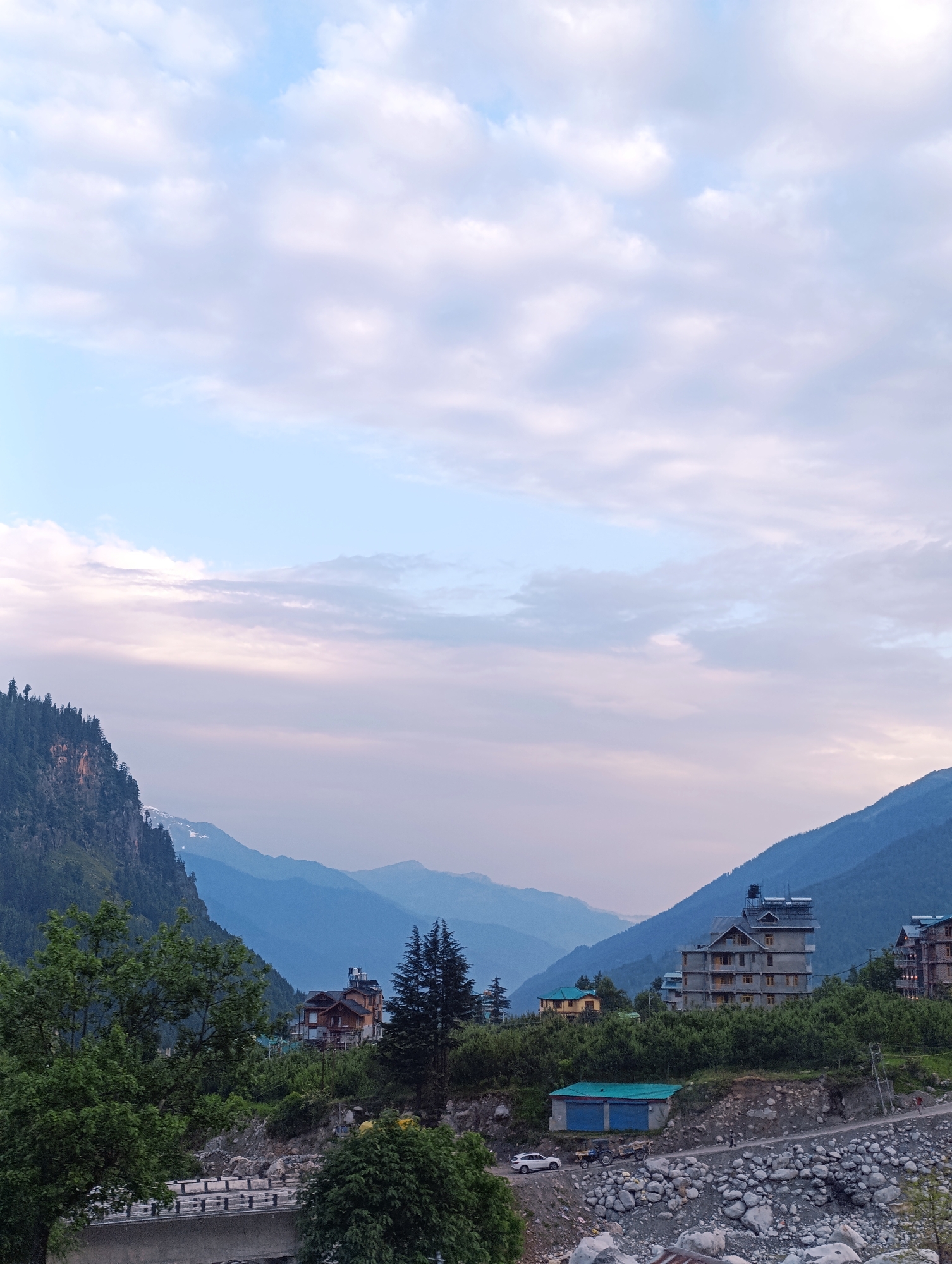 In Palchan, India, a wide shot captures a valley landscape at dusk. The sky is a gradient of soft blues and pinks, filled with scattered white clouds. Majestic, layered mountains dominate the background, fading into a hazy blue. In the foreground, a dense forest of green trees covers the slopes. A few houses are nestled amongst the trees and on the mountainside. A riverbed, filled with large rocks, runs through the lower part of the image. A road with a few vehicles, including a white car and a tractor, is visible. A bridge crosses the river in the left foreground. The overall impression is one of natural beauty and tranquility.