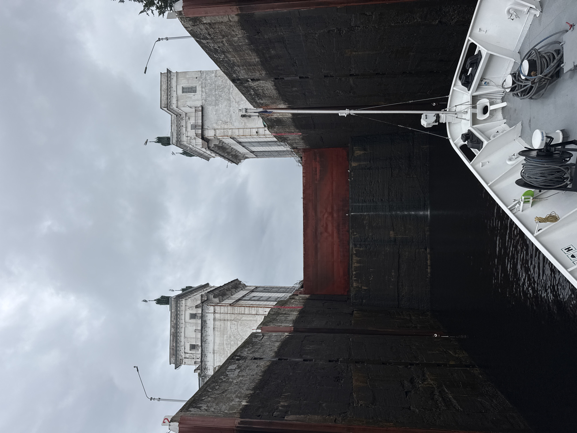 The image shows a cargo ship entering a lock on a cloudy day in Podporozhye, Russia. The lock gates are massive, dark structures, with a red band visible on the lower portion of the right gate. The ship, painted white, is partially visible on the right side of the frame, its bow curving towards the lock. The deck of the ship is equipped with various ropes and equipment, including a winch with coiled rope and a cylindrical white object that appears to be a beacon or sensor.
In the background, on the left side of the frame, are imposing, weathered stone buildings with arched windows and tiered rooftops. These structures suggest a historical or industrial setting. Green-capped spires rise from the rooftops of these buildings. The sky is overcast with heavy grey clouds, creating a muted light and a somber atmosphere. The water in the lock is dark and still, reflecting the overcast sky. The overall scene depicts the process of a ship navigating a lock system, likely for a change in water level. No people are visible in the image. There is no visible text in the image.