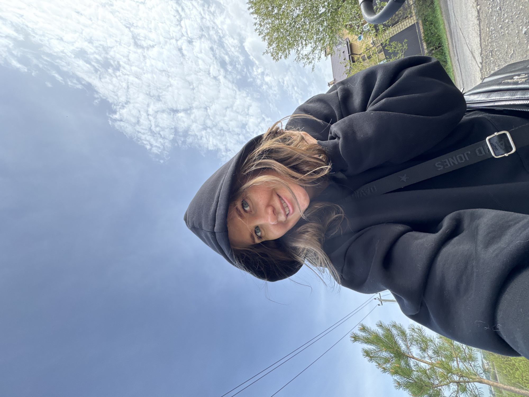 A selfie taken at an upward angle shows a woman in a black hooded sweatshirt, with a "David Jones" bag strap visible over her shoulder. She is smiling slightly, looking directly at the camera. The background includes a blue sky with scattered clouds, trees, and a residential street. The location is Горный, Russia.