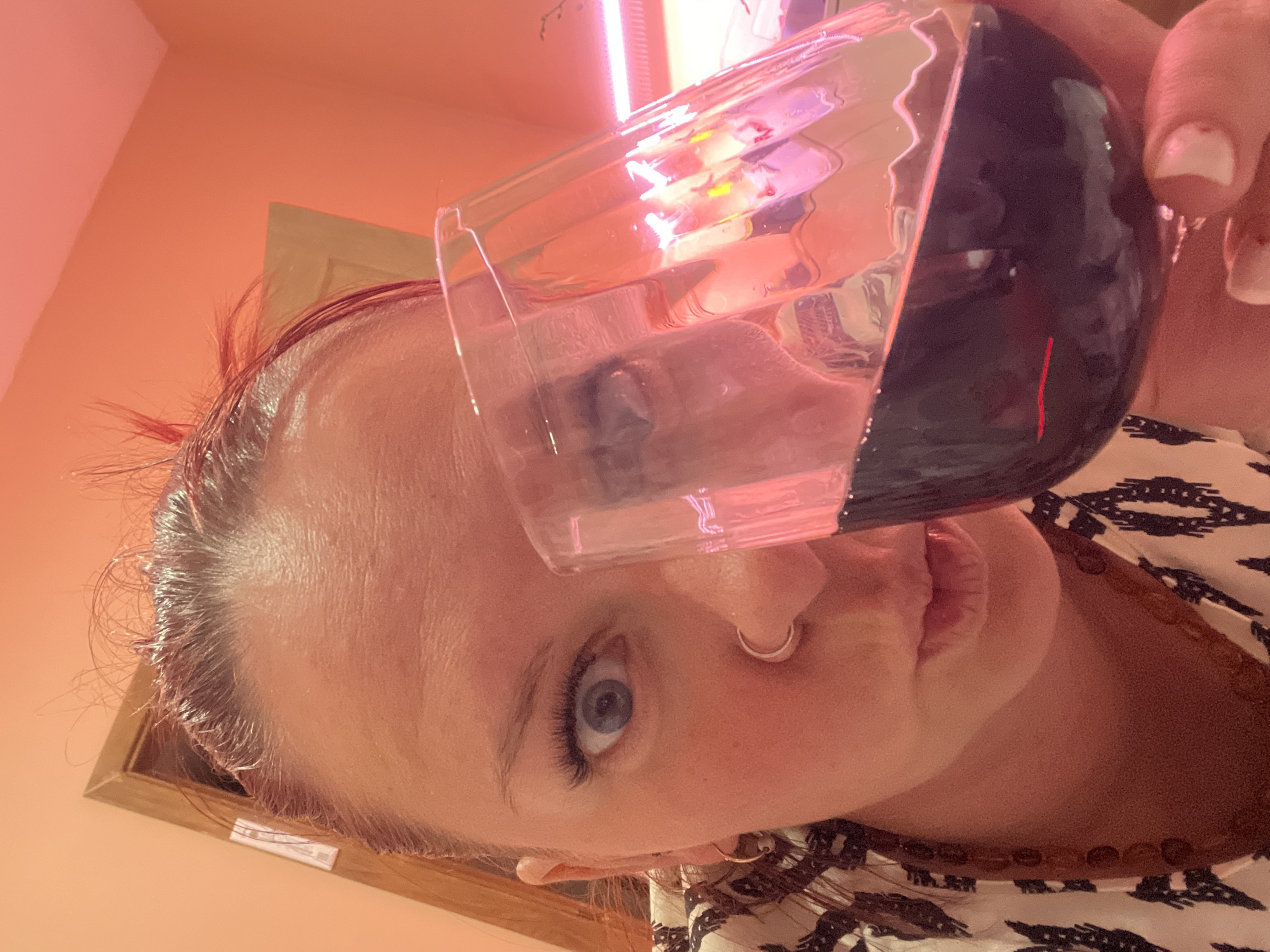 A person is holding a glass of dark liquid in front of their face. The person has fair skin, blue eyes, and a nose ring. They are wearing a patterned shirt and a necklace. The background is a light orange color with a rectangular frame on the left side. There is also a bright light source at the top of the image. The image is rotated, so the objects are positioned at an angle. The location in Chaikovsky, Russia, is not determinable from the visual information provided.