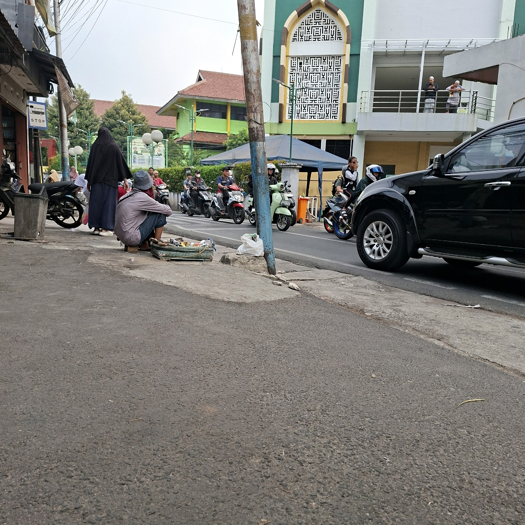 A medium-angle, eye-level shot captures a street scene in an urban environment. In the foreground, a person in a blue shirt and gray hat crouches on the pavement, sorting through items laid out in a green tray on the ground. Behind them, a black SUV is parked, its front wheel visible. To the left, a dark motorcycle is parked next to a trash bin.
Further into the scene, several people are on motorcycles, either riding or stopped. A woman in a black hijab and a dark blue skirt walks away from the camera. In the background, a green and white building with Arabic calligraphy on its facade stands prominently, with a balcony occupied by two men. Other buildings and trees are visible, suggesting a bustling street with pedestrian and vehicular activity. The sky is overcast.