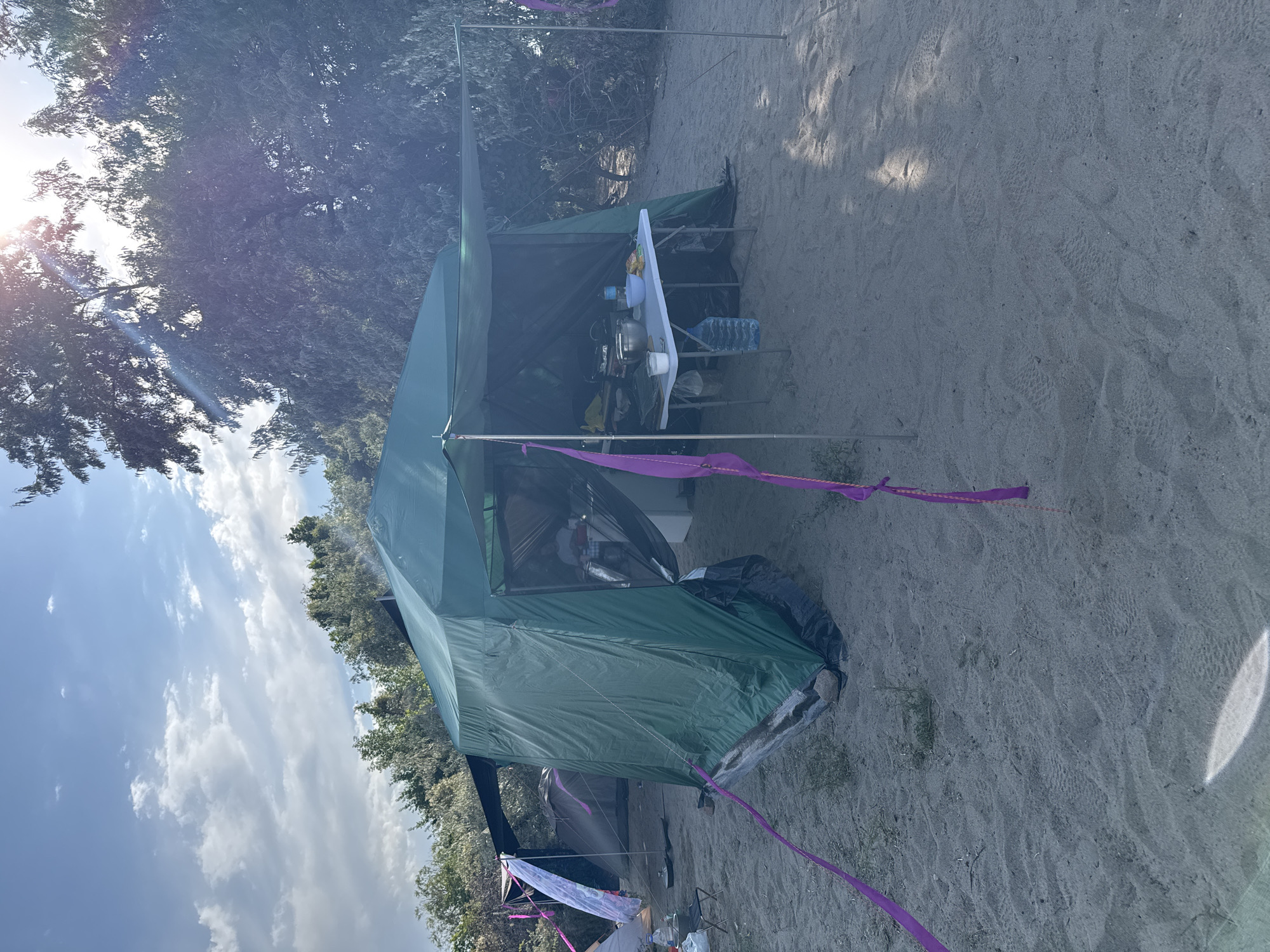 The image depicts a campsite on a sandy area, likely near a body of water given the presence of trees and sparse vegetation in the background. The main subject is a large, green tent, set up with an attached canopy that extends over a folding table. The table is laden with various items, including a large metal bowl, a blue water bottle, and food items. A person can be seen seated at the table, though their features are not clearly discernible. Adjacent to the main tent, another tent is visible, partially obscured by the larger structure. Purple ribbons are tied to tent poles and also draped from the canopy, suggesting some form of decoration or demarcation. The ground is composed of fine sand, with some footprints visible. Sunlight is filtering through the trees and clouds, indicating it is daytime. The scene is peaceful and appears to be a recreational camping setup. The location is identified as Basbeshik, Kyrgyzstan.