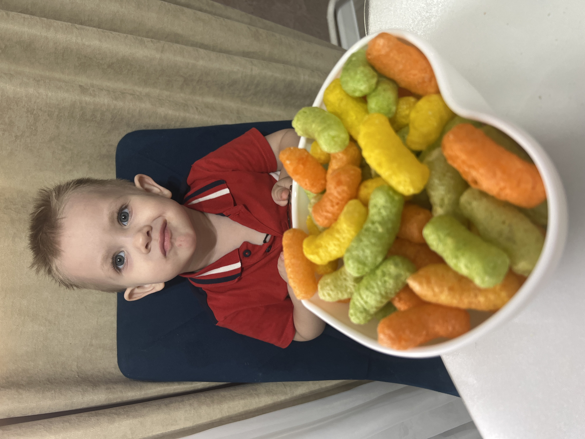 A toddler with fair skin, blond hair, and blue eyes is reclining in a dark blue chair. He is wearing a red polo shirt. The boy holds a white, heart-shaped bowl filled with colorful puffed corn snacks in orange, yellow, and green. The bowl is positioned above the boy, held over a white surface. The backdrop consists of a beige curtain and a white curtain. It is unlikely that the specific location is Марковский, Russia, is discernible from the image alone.