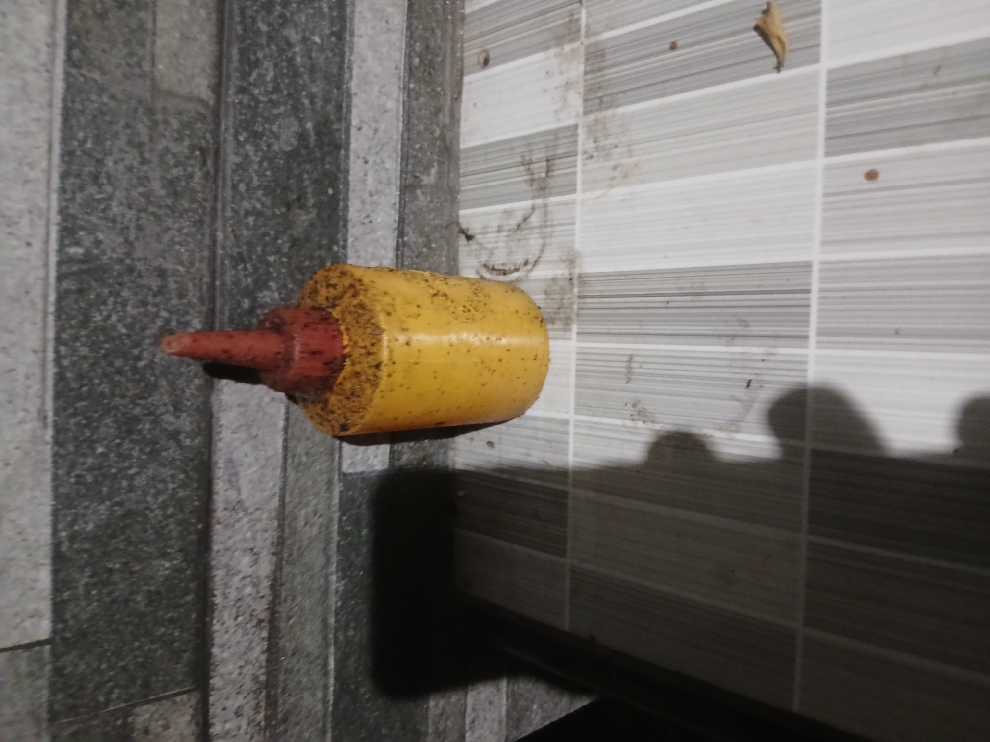 A yellow cylindrical object with a red conical cap sits on a tiled surface. The object appears to be some sort of dispenser or container, with small dark specks adhered to its surface, particularly around the top. A dark shadow is cast to the left of the object, suggesting a light source from the right. The background consists of a wall with horizontally oriented gray tiles. The tiled surface the object rests on is light-colored with a pattern of alternating white and gray stripes. A single dried leaf is visible on the tiled surface to the right of the object. The scene is located in Sekarputih, Indonesia.