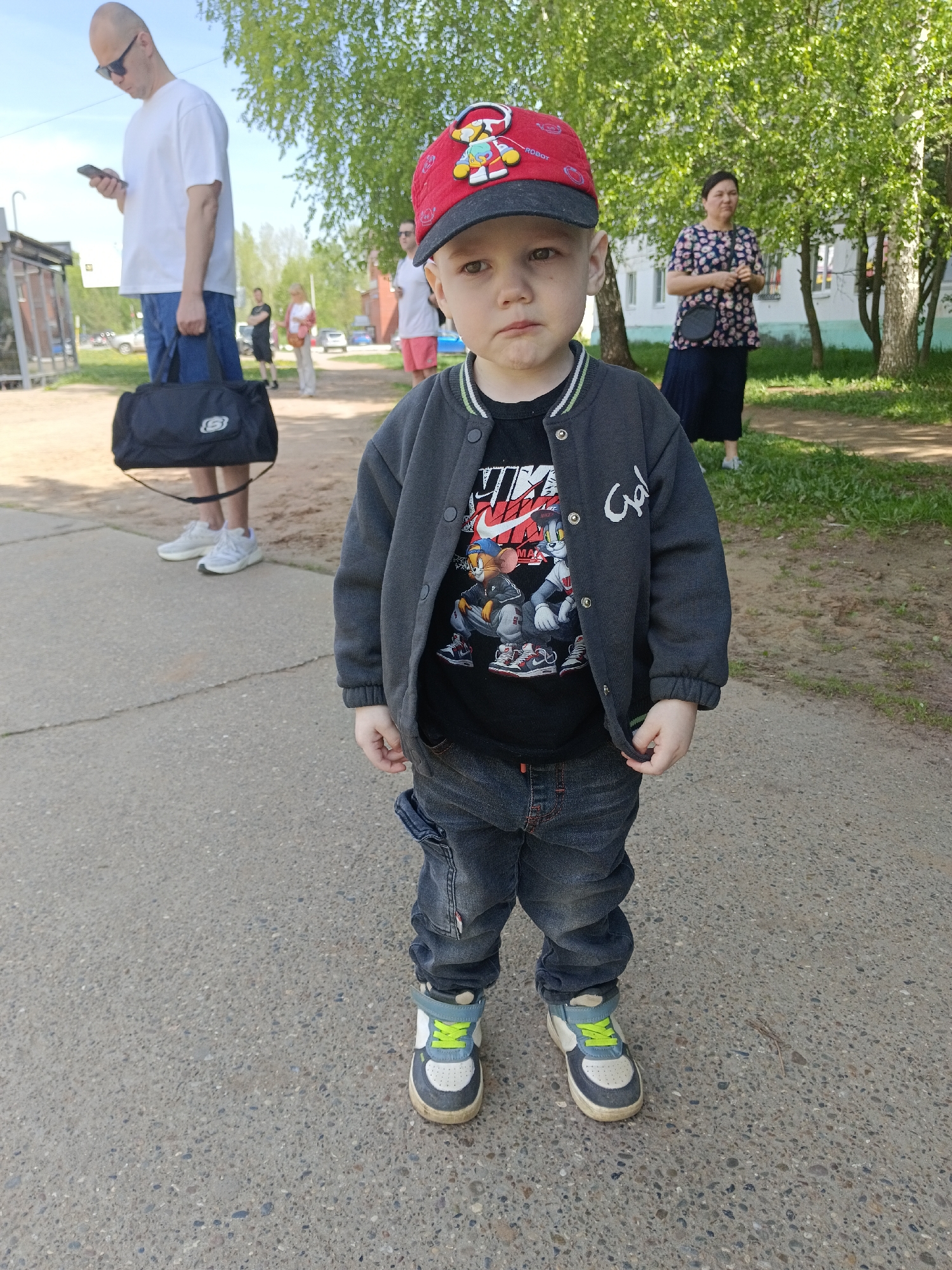 A young boy stands on a concrete surface, facing the camera. He wears a red cap, a dark jacket over a printed t-shirt, dark jeans, and two-tone sneakers with green laces. In the background, a man in a white t-shirt and blue shorts holds a black duffel bag while looking at a cell phone. Other people stand in the background, including a woman in a patterned blouse and dark skirt. Green trees and some buildings are visible. The photo was taken in the city of Chaikovsky, Russia.