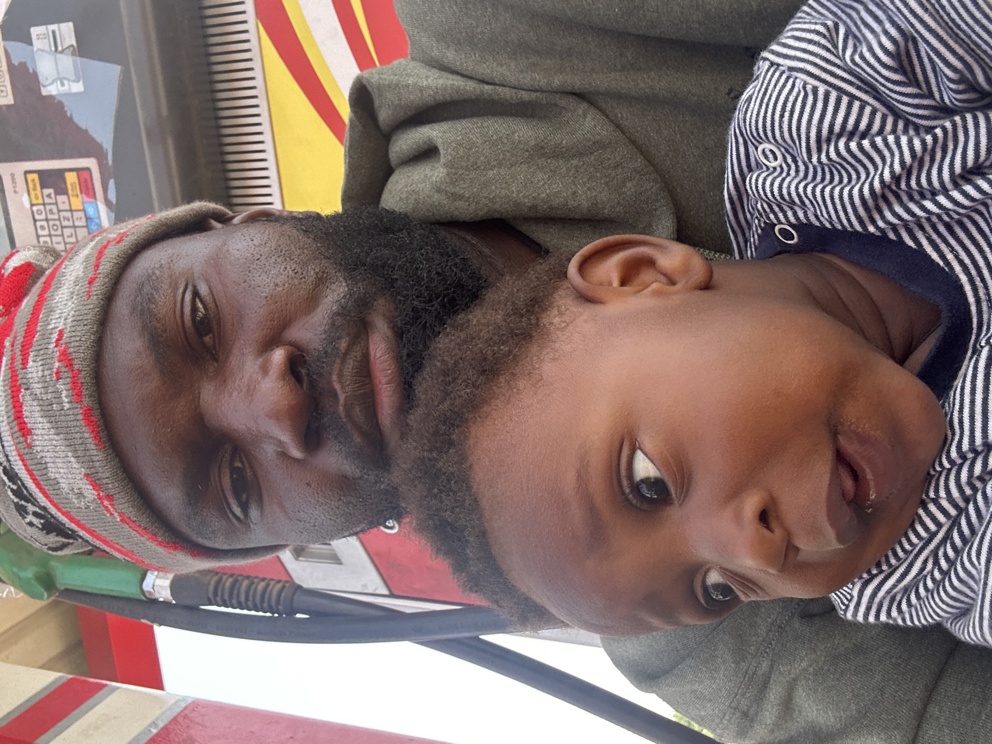 The media file shows a close-up, rotated image of a man and a baby. The man, on the left, has dark skin and is wearing a gray hoodie and a beanie with red and gray stripes. He has a beard and appears to be looking towards the camera with one eye visible. The baby, on the right, also has dark skin and is wearing a blue and white striped onesie. The baby's face is turned, and one eye is visible, with its mouth slightly open and tongue sticking out. The background is blurred but shows some colorful vertical stripes, possibly part of a structure or a sign. A green gas nozzle with a black hose is visible in the foreground, extending from the bottom left corner upwards, suggesting the scene might be at a gas station. The lighting appears to be natural, indicating it could be daytime. The overall impression is an intimate moment captured between the man and the baby. There is no visible text that can be clearly deciphered. The location context is Gura Top, Nigeria.