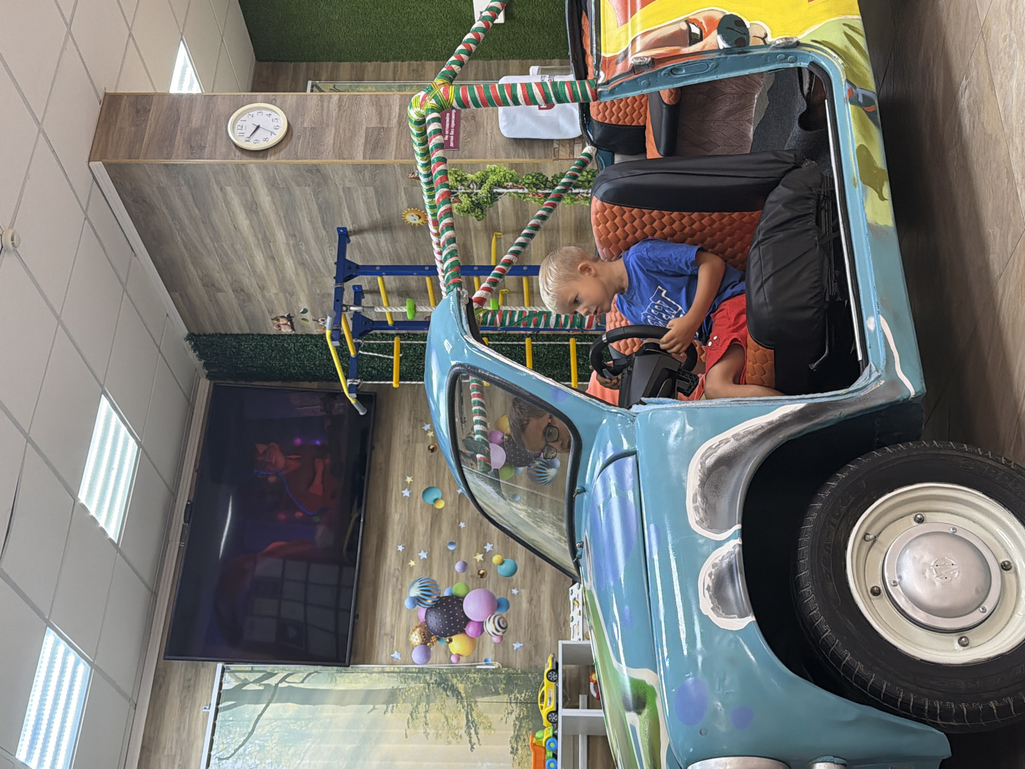 A young boy with blonde hair, wearing a blue t-shirt and red shorts, sits in the driver's seat of a light blue toy car with a colorful, cartoonish exterior, gripping the steering wheel. He is looking to his right. Another child with dark hair and glasses is visible in the passenger seat, also looking to the right. The car is positioned indoors, on a tiled floor. In the background, a large television screen displays an animated image. There is a wooden-paneled wall with a clock on it, and a green wall with a hedge-like texture. A climbing frame structure is visible behind the toy car. This scene appears to be in a play area or entertainment center, possibly located in Sochi, Russia, given the context of the surrounding image details.