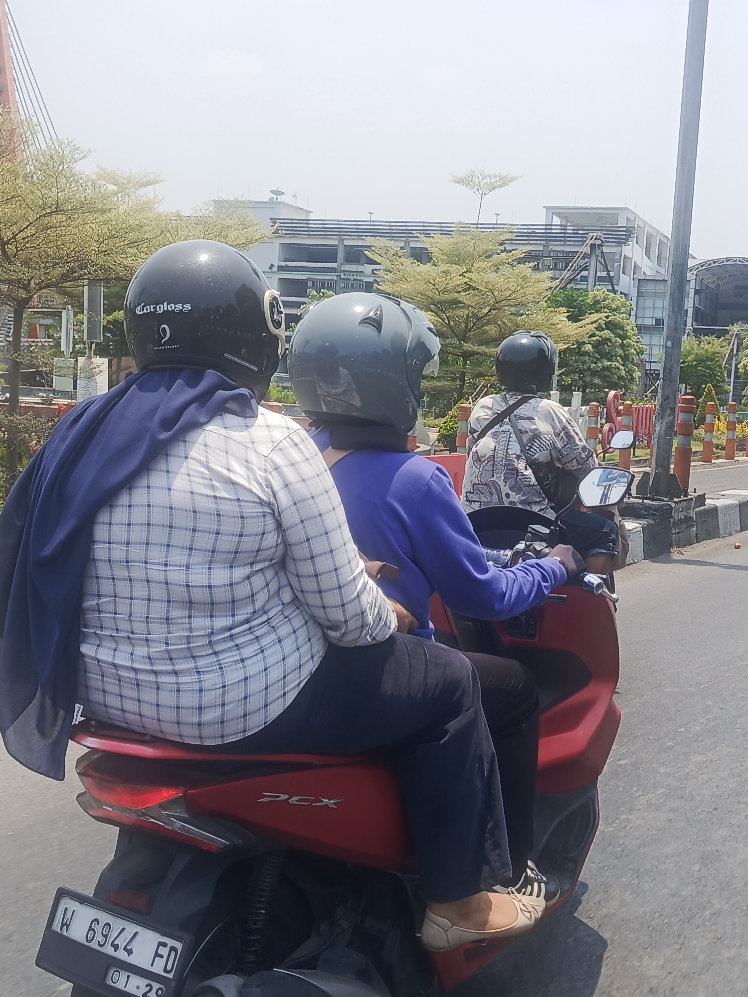 Three individuals are riding on a red scooter in the city of Surabaya, Indonesia. The person in the front, wearing a blue long-sleeved shirt and dark pants, is operating the scooter. Behind them, a second person is wearing a black helmet with "Cargloss" branding and a blue shawl draped over their shoulders, paired with a white and blue checkered long-sleeved shirt and dark pants. The third person, at the rear, is wearing a black helmet and a patterned shirt. The scooter is branded with "PCX". The background shows a clear sky, trees with light-colored foliage, and a multi-story building. Orange traffic cones and a gray concrete barrier line the right side of the road, indicating an urban setting. The scooter's license plate reads "W 6944 FD" with the date "01-29" below it. The overall scene depicts transportation within a city.