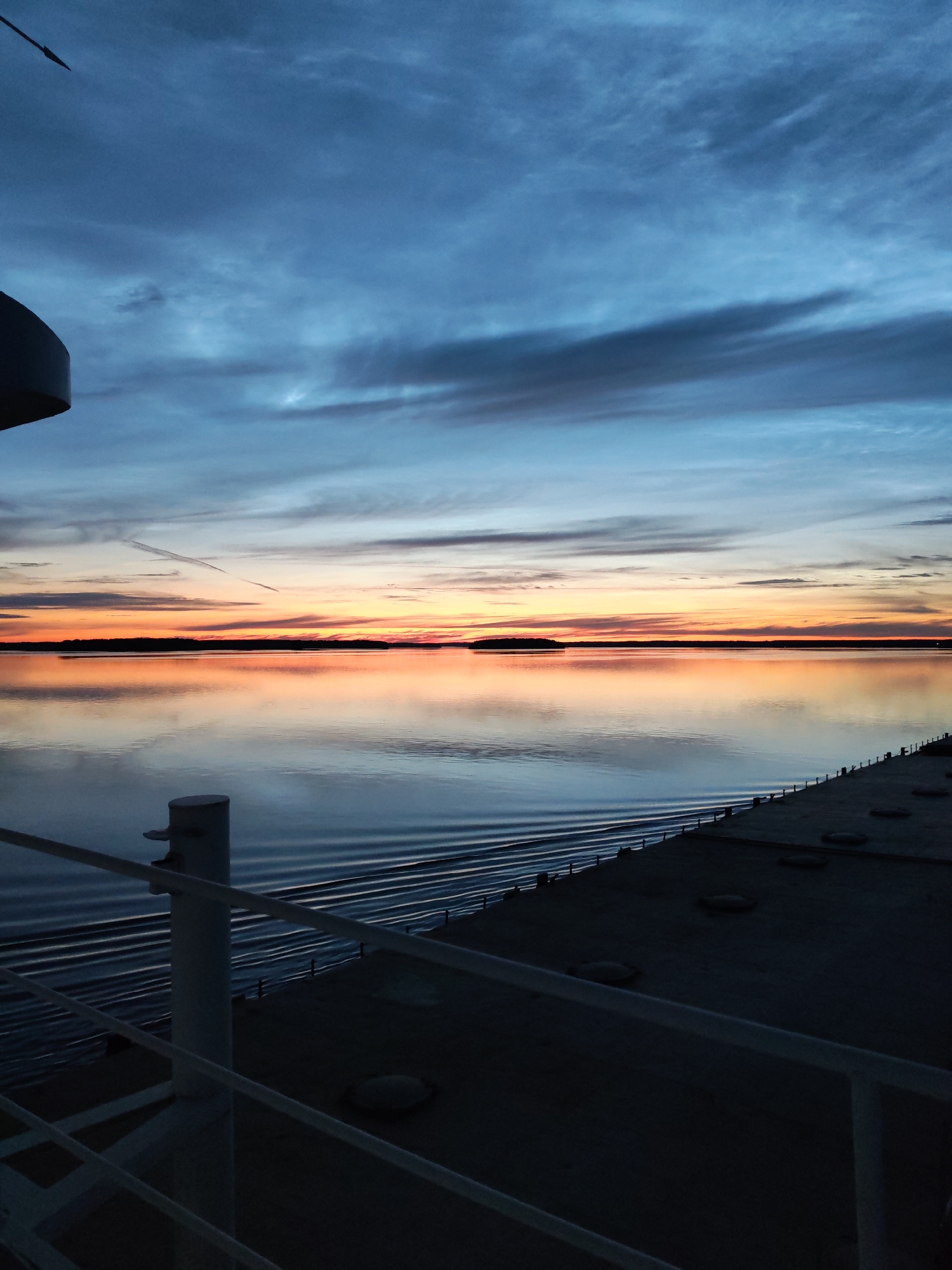 The image depicts a sunset over a body of water, possibly a lake or sea, viewed from the deck of a vessel. The sky is a gradient of deep blue at the top, transitioning to lighter blues and then to vibrant oranges and pinks near the horizon, with scattered clouds. The water is calm and reflects the colors of the sky, creating a mirror-like effect. A distant shoreline is visible as a dark silhouette against the colorful sky. In the foreground, the deck of the vessel is visible, with railings and a bollard. The scene appears to be tranquil and atmospheric, capturing the natural beauty of the sunset. There are no people visible in the image. The location is identified as Malinukha, Russia.