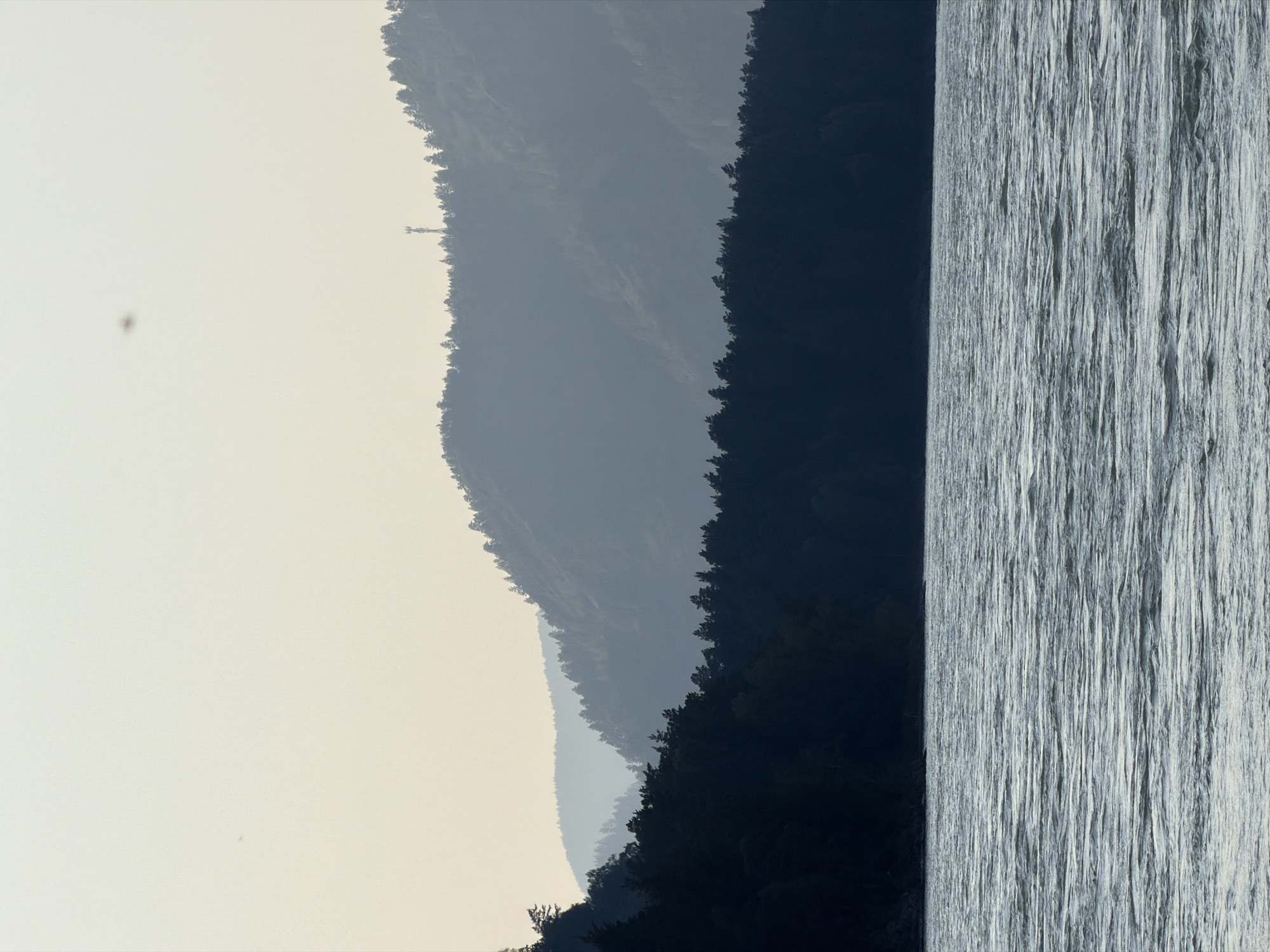 The image depicts a serene landscape of mountains, a forest, and a body of water. In the foreground, a wide river with rippling water flows, reflecting the subdued light. The midground features a dense, dark forest covering the lower slopes of rolling hills. Further in the background, layers of hazy, blue-hued mountains recede into the distance, their peaks softened by the atmospheric perspective. Atop one of the distant mountain ridges, a solitary cell tower is visible, a man-made structure contrasting with the natural environment. The sky is a pale, overcast grey, with a faint reddish-orange object suspended in the upper portion of the frame, possibly a distant balloon or drone. The scene evokes a sense of tranquility and vastness, characteristic of natural landscapes. The location is identified as Anos, Russia.
