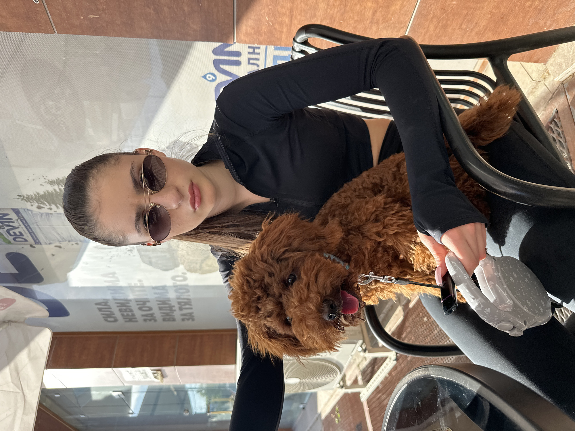 A young woman and her dog are pictured in an outdoor setting, likely a cafe or restaurant patio in Plovdiv, Bulgaria, on a sunny day. The image is oriented vertically (rotated 90 degrees clockwise from the original view).
The woman, with dark hair pulled back, wears round, gold-rimmed sunglasses, a black long-sleeved athletic top, and black leggings. She holds a grey retractable dog leash adorned with paw print patterns. Her expression is neutral. A brown, curly-haired dog, resembling a poodle or poodle mix, is seated close to her, possibly on her lap. The dog has its tongue slightly out, suggesting warmth or relaxation, and wears a collar connected to the leash.
They are seated on black metal chairs with horizontal slats. A dark, possibly reflective table surface is partially visible in the foreground. The background consists of a storefront window or glass partition, which reflects parts of the urban environment. Bright sunlight creates clear shadows, indicating a bright, clear day.
Text visible on the glass or a sign in the background includes Cyrillic script. Decipherable words are "DEVIN" (a Bulgarian bottled water brand), "СИЛА" (Strength), "НЕВИДИМА" (Invisible), "ЗА ОЧИТЕ" (For the eyes), "ВИДИМА" (Visible), and "ЗА ТЯЛОТО." (For the body.). A logo resembling a water droplet or an eye symbol, along with the number "6," is also present near the "DEVIN" text. The scene captures a tranquil moment of companionship between the woman and her pet outdoors.