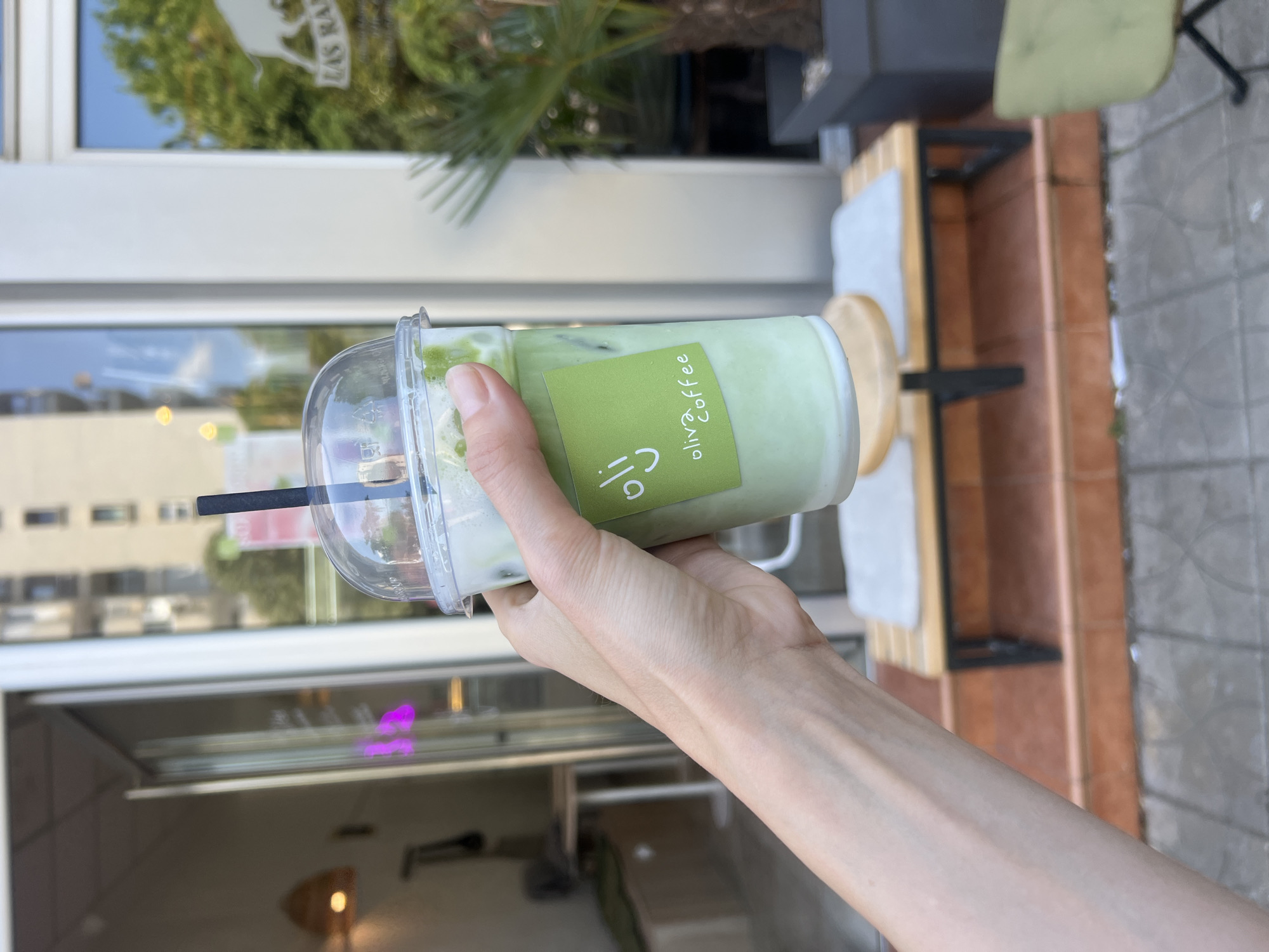 A light-skinned hand is holding a plastic cup of matcha latte with a black straw and a green label reading "Olive Coffee." The cup contains ice. The background shows a building with a glass storefront reflecting the city. Outside the storefront, a black metal table is seen with a chair. A palm tree is also visible. The location is likely Bar, Montenegro, based on the "Olive Coffee" brand, which is locally known.