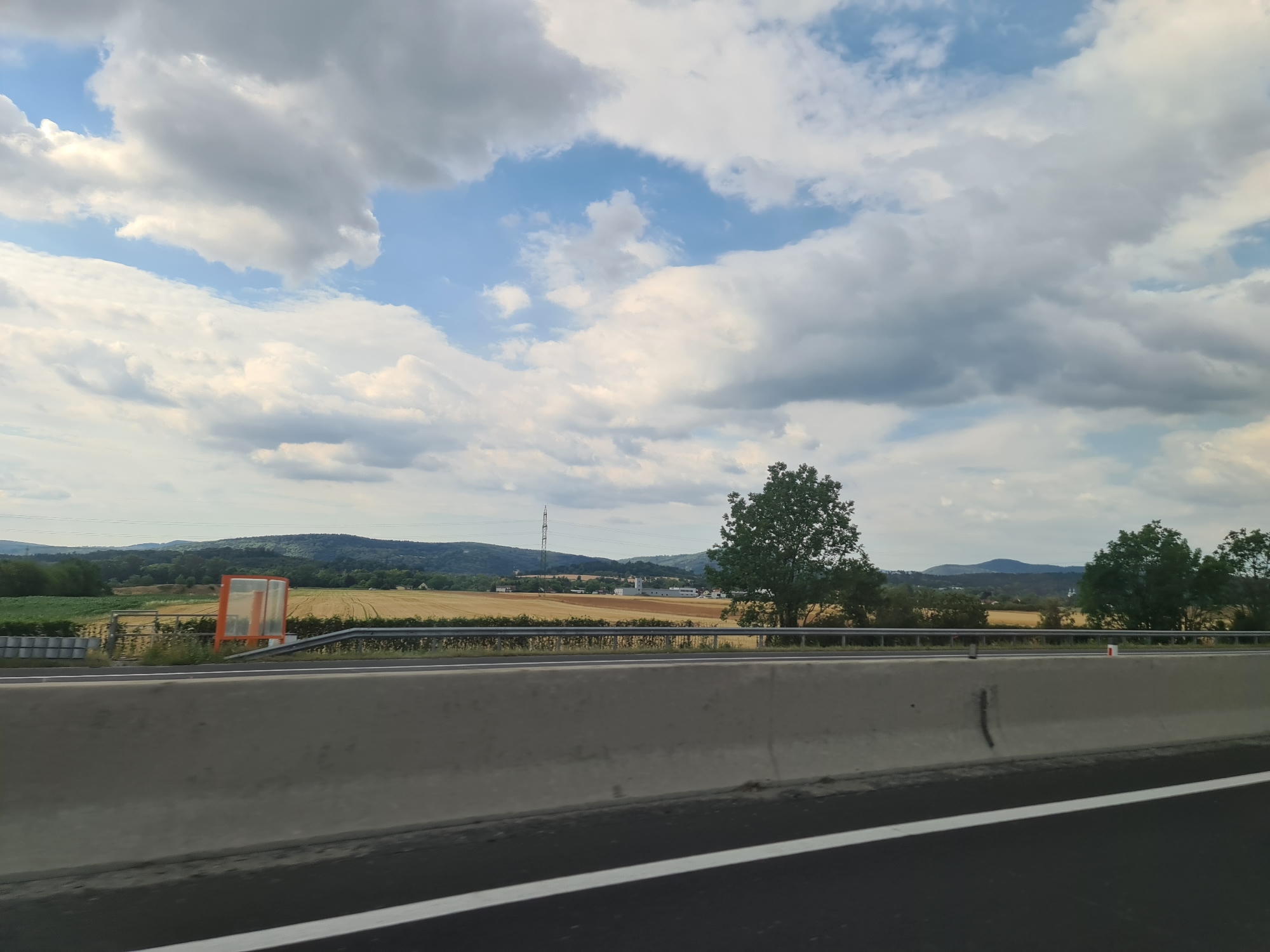 The image depicts a view from a highway in Föhrenau, Austria, looking towards rolling hills under a partly cloudy sky. A concrete barrier is in the foreground, with a white line marking the edge of the asphalt road. Beyond the barrier, there is a metal fence. In the middle ground, fields of golden crops, likely harvested wheat or barley, stretch across the landscape. A single transmission tower is visible in the distance, along with scattered trees and buildings. The sky is filled with a mix of white and gray clouds, with patches of blue visible. The overall scene suggests a rural setting viewed from a passing vehicle. There are no people or animals visible in the image.