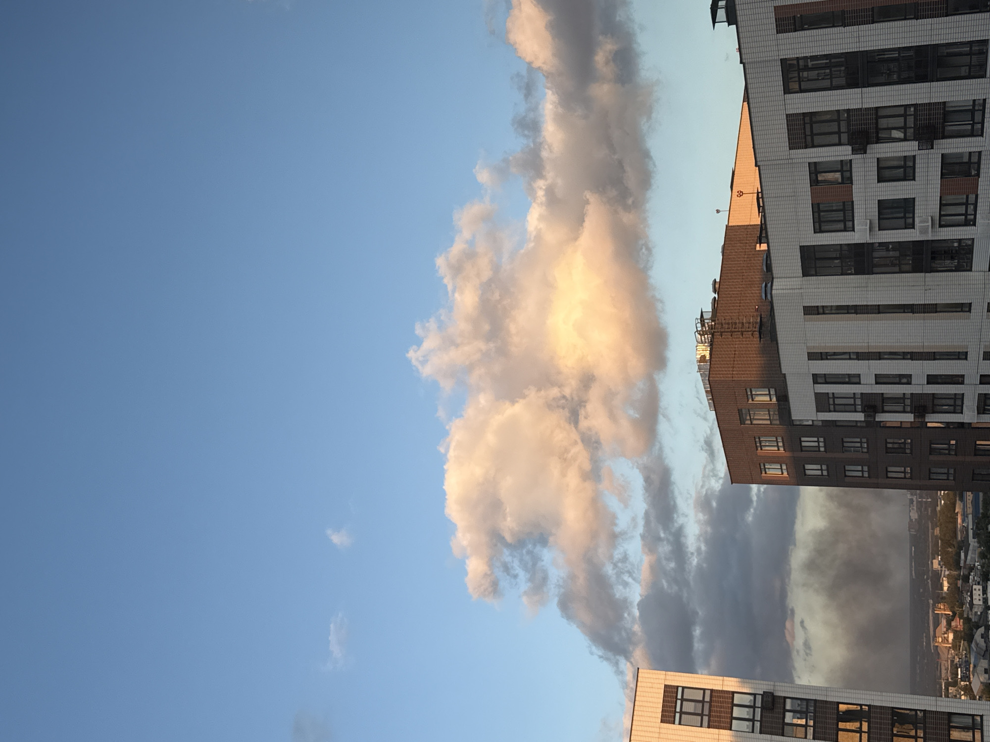The image shows a view of the city of Lyubertsy, Russia, on a clear day with scattered clouds. In the foreground, there are several modern apartment buildings. The sky is blue with some pink and orange hues from the setting sun. In the middle ground, a plume of dark smoke rises from what appears to be a fire in the distance, obscuring the cityscape.