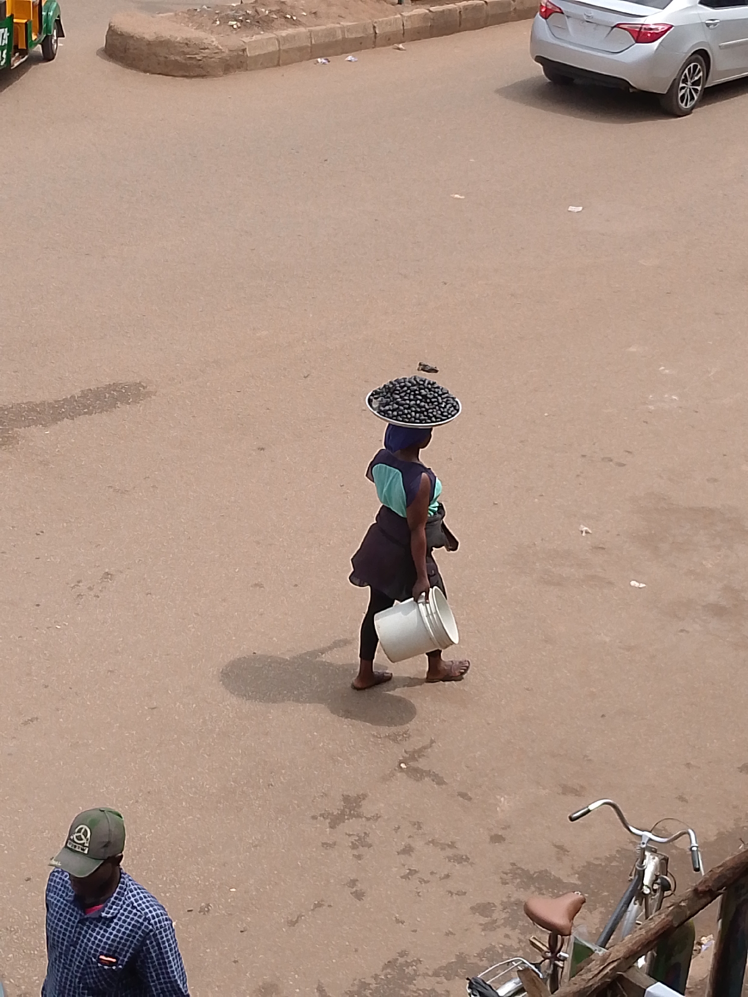 In Bukuru, Nigeria, a woman walks along a paved road, carrying a large platter of dark, spherical items, likely produce, balanced on her head. She is wearing a blue and green top and dark pants. In her right hand, she holds a white bucket. To her right, the shadow of a bicycle is visible, with the actual bicycle parked near a wooden barrier. Further down the road, a man wearing a plaid shirt and a green cap is walking away from the camera. A silver car is parked in the background on the right side of the image, and a green and yellow vehicle is visible on the far left. The ground is a dusty, light brown color, with some scattered debris.