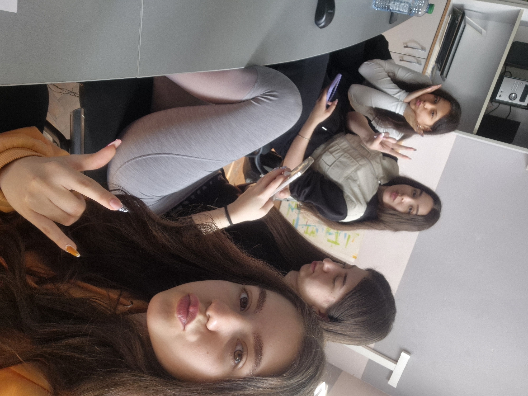 Four young women are indoors, presumably in Pavlikeni, Bulgaria. The photo appears to be a selfie taken by one of the women. The woman closest to the camera has long brown hair and is wearing an orange sweater. She's making a hand gesture. Behind her is another young woman with brown hair. Two other young women are visible in the background, one holding a cell phone and another making a peace sign. A desk, water bottle, and drawers are visible in the background.