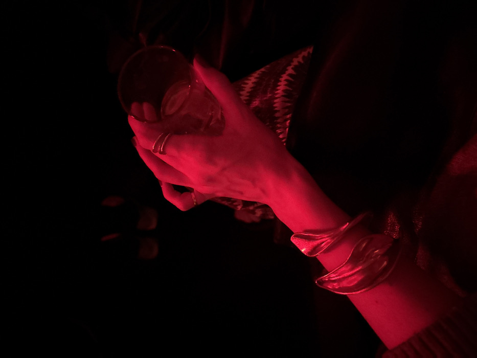 A close-up photograph, heavily illuminated by red light, captures a human left hand holding a clear, cylindrical glass containing a beverage and ice cubes. The hand wears dark nail polish, two rings on the index finger, and one ring on the ring finger. A large, metallic, gold-toned cuff bracelet with an abstract, folded design is prominently displayed on the wrist. Beneath the hand, a dark textile with a visible zigzag pattern is present, resting on a dark, smooth surface, possibly leather. The background is predominantly black and out of focus, with indistinct reddish light sources in the upper portion. No specific landmarks or architectural features are discernible to identify a precise location within the city of New York, United States of America. The scene suggests an indoor social environment, such as a bar or lounge.