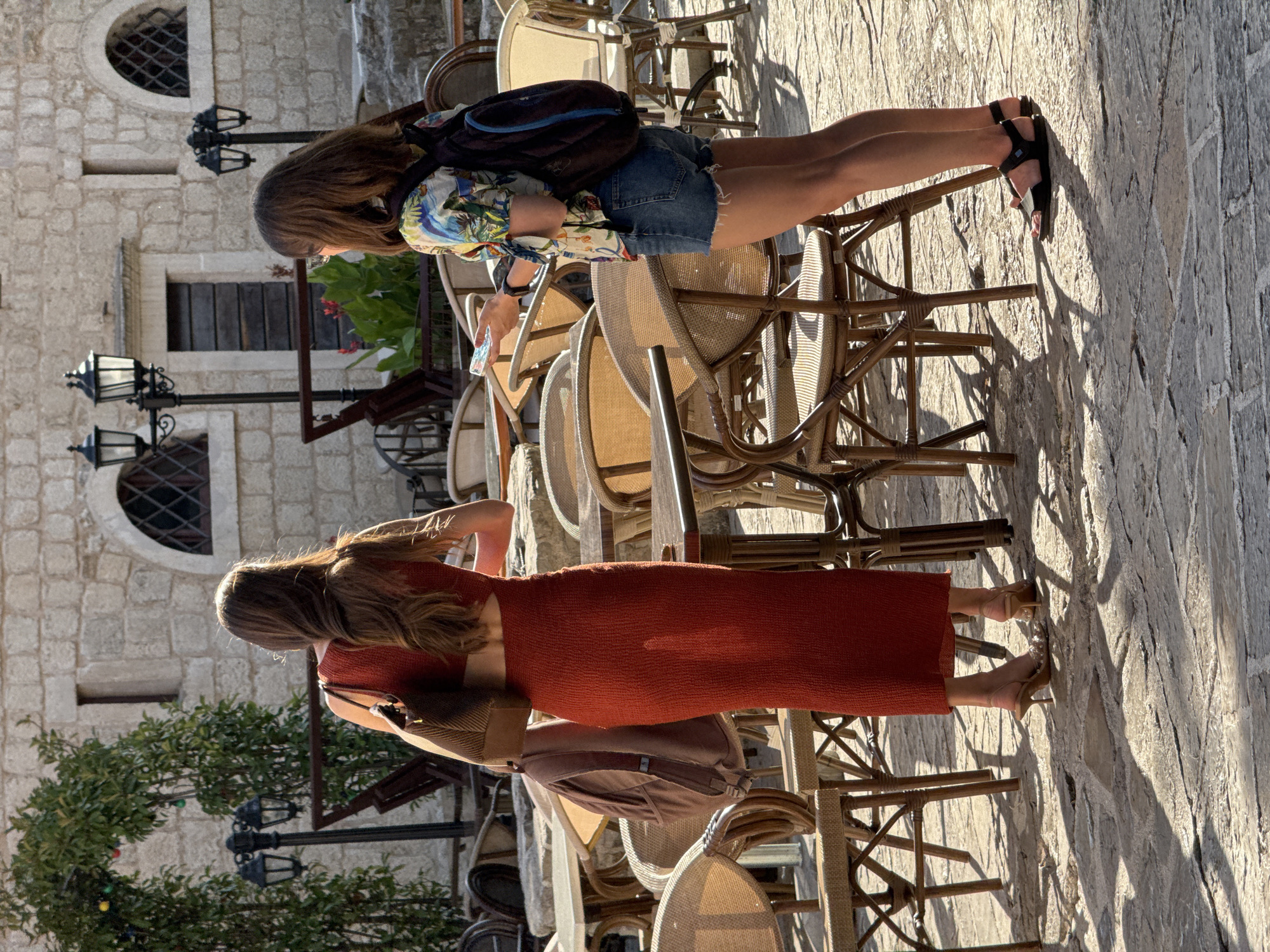 The image depicts two women standing in an outdoor seating area of a cafe in the old town of Budva, Montenegro. The woman on the left, seen from behind, wears a long, form-fitting orange dress and carries a brown shoulder bag. Her long brown hair is down her back. The woman on the right, also viewed from behind, wears a short-sleeved, colorful patterned shirt, denim shorts, and a backpack. She is holding a smartphone in her right hand. They are surrounded by numerous empty wicker chairs and tables, with stone buildings and an archway covered in greenery in the background. The ground is paved with cobblestones, and the scene is illuminated by sunlight, casting distinct shadows.