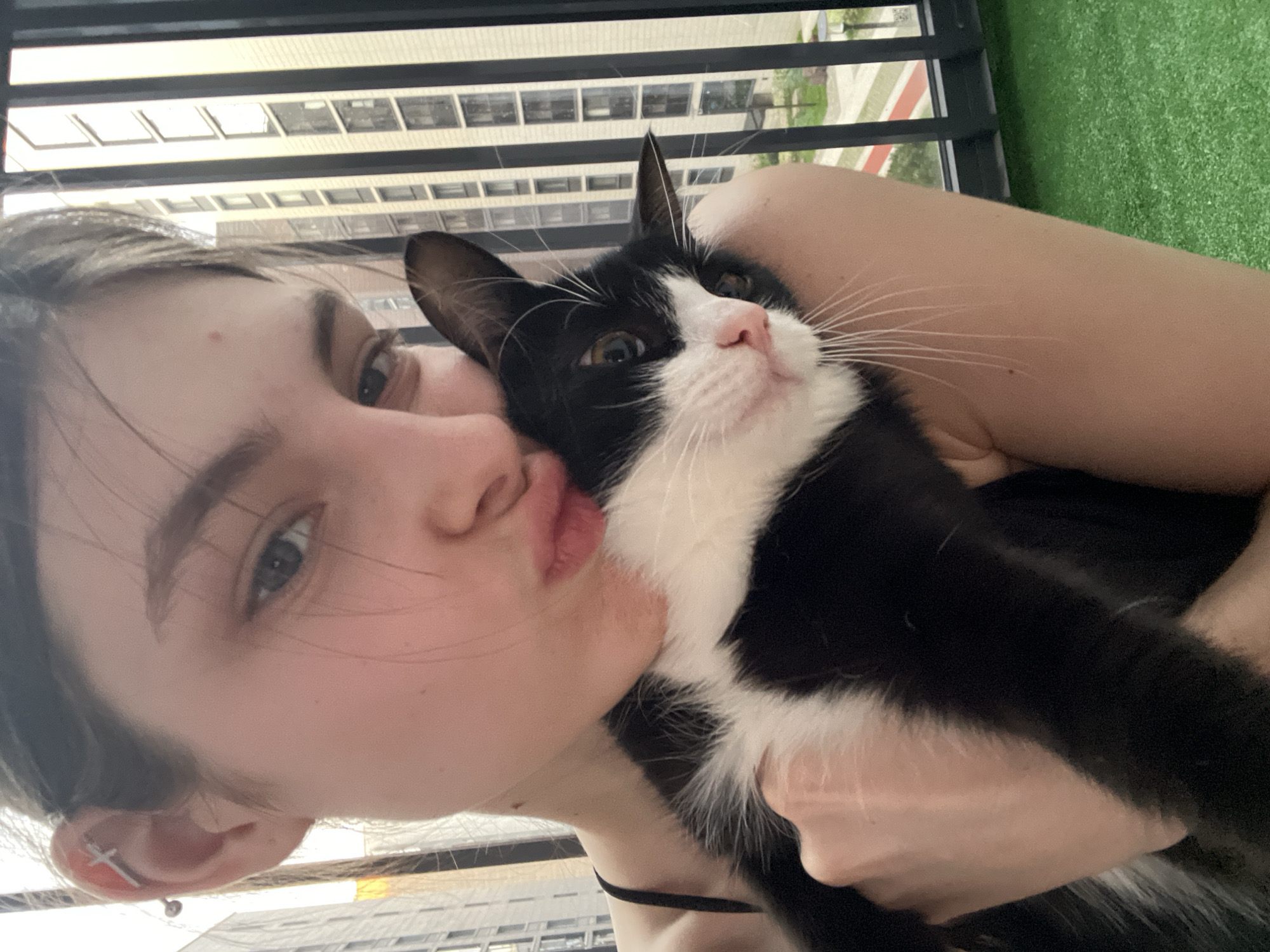 A young woman is holding a black and white cat. She is kissing its head. The woman has fair skin, light eyes, and dark hair. She is wearing a black top. The cat has a pink nose and long white whiskers. They are on a balcony. The background shows buildings, green turf on the balcony and a metal railing. The city cannot be verified.