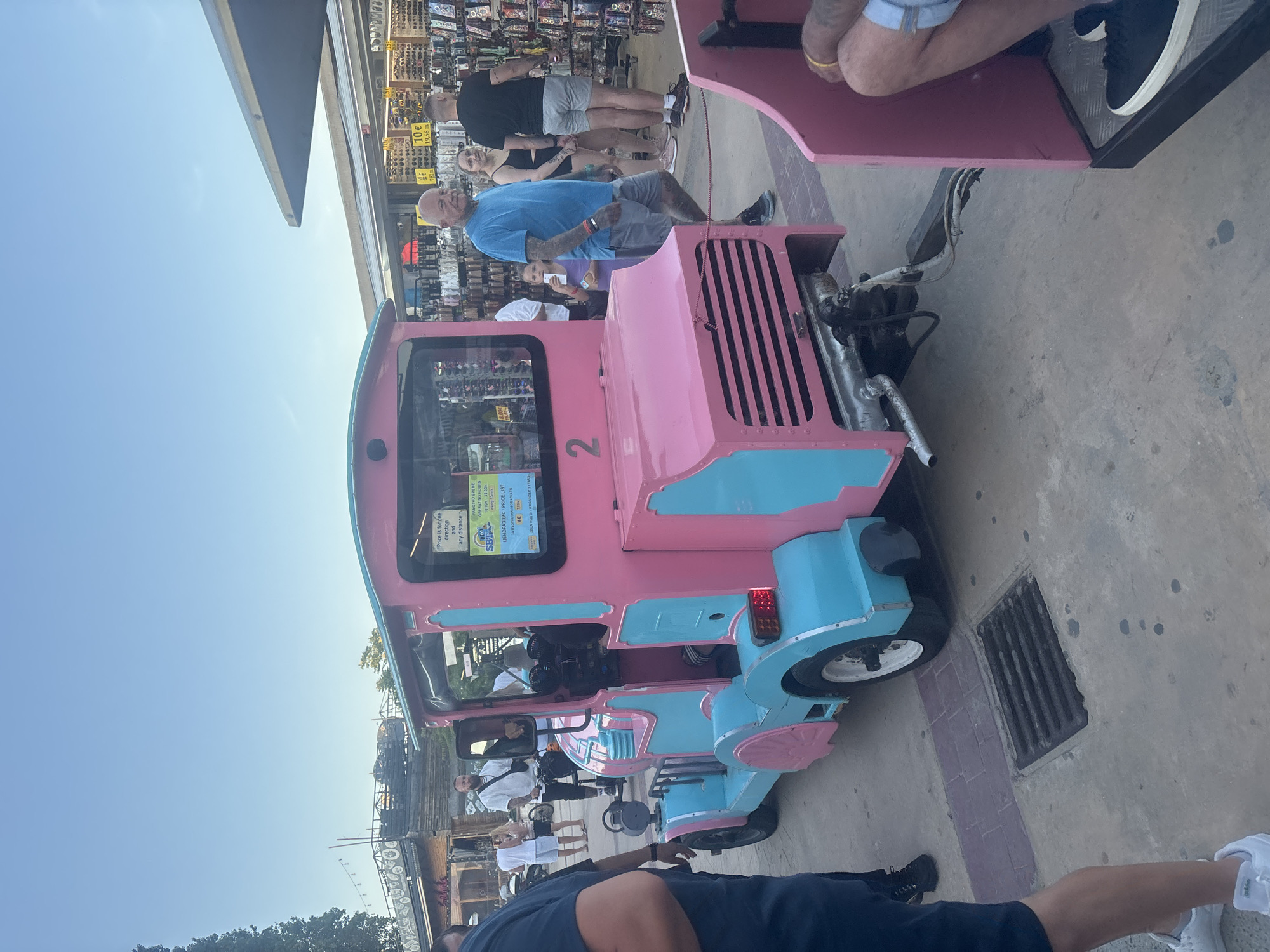 The image shows a pink and turquoise train-like vehicle with the number 2 on its side, parked on a paved area with a grate in the foreground. The vehicle appears to be a tourist attraction, possibly a road train for rides. In the background, there are several people. To the right of the train, a man in a blue t-shirt and shorts stands looking towards the camera, with a woman behind him and another man in a black t-shirt and grey shorts further behind. Behind them, there are market stalls selling various items, including sunglasses. To the left of the train, a man in a blue shirt and black shorts stands with his back to the camera. The overall scene suggests a public, possibly tourist area in a city. Based on the signs in the image, which include Cyrillic script and prices in Euros, and the architecture visible in the background, this scene is located in Nesebar, Bulgaria, likely on the Old Town peninsula. The sky is clear and blue, indicating daytime.