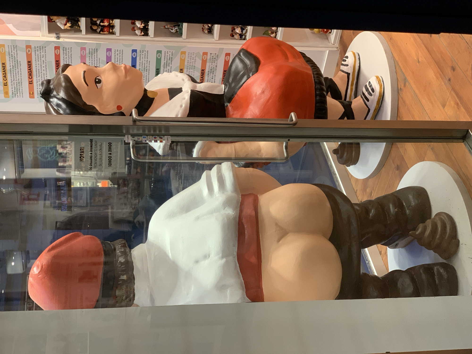 The image shows two statues of "El Caganer" figures, a traditional Catalan Christmas character, displayed in a storefront window, likely in Barcelona, Spain. The top figure is a man wearing a red and black hat, a white shirt, a red sash, and dark trousers. He is depicted in a squatting position. Below, a similar figure is shown with the red hat, white shirt, and red sash. He's also squatting with his buttocks exposed and a small pile of excrement depicted behind him. The statues are on a white base. The window also shows a sign with the words "El Caganer" and "Horari" (schedule), indicating store hours. The sign is in multiple languages. The reflections on the glass make it difficult to see the background clearly, but the location is likely a shop in Barcelona.