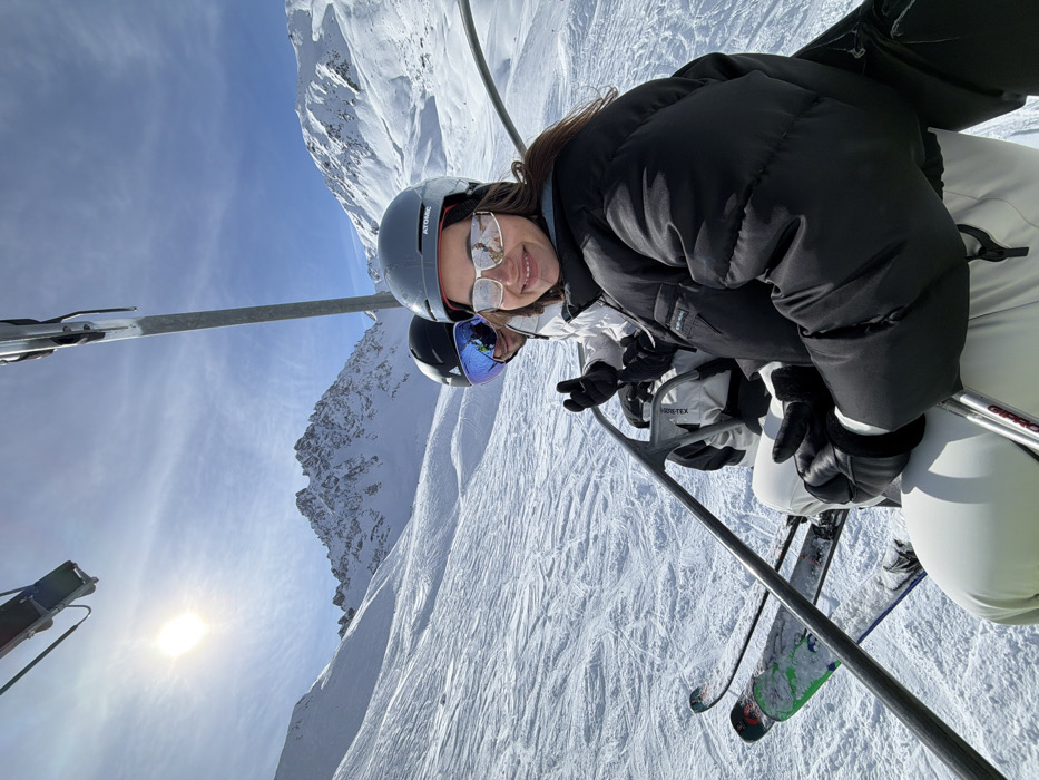 Two individuals are observed ascending a snow-covered mountain on a ski chairlift in Courchevel, France. The foreground features a female with dark hair, wearing a grey Atomic-branded ski helmet, large reflective silver sunglasses, a black puffer jacket, black gloves, and light-colored ski pants. She is smiling and facing the camera. Behind and to her left, a second person is partially visible, wearing a black ski helmet, blue mirrored ski goggles, a white Gore-Tex branded jacket, and black gloves. Below them, a pair of skis with green and black markings hangs from the chairlift. The environment consists of vast snow-covered slopes with visible ski tracks, prominent rocky mountain peaks in the background, and a bright sun in a clear blue sky, indicating sunny weather.