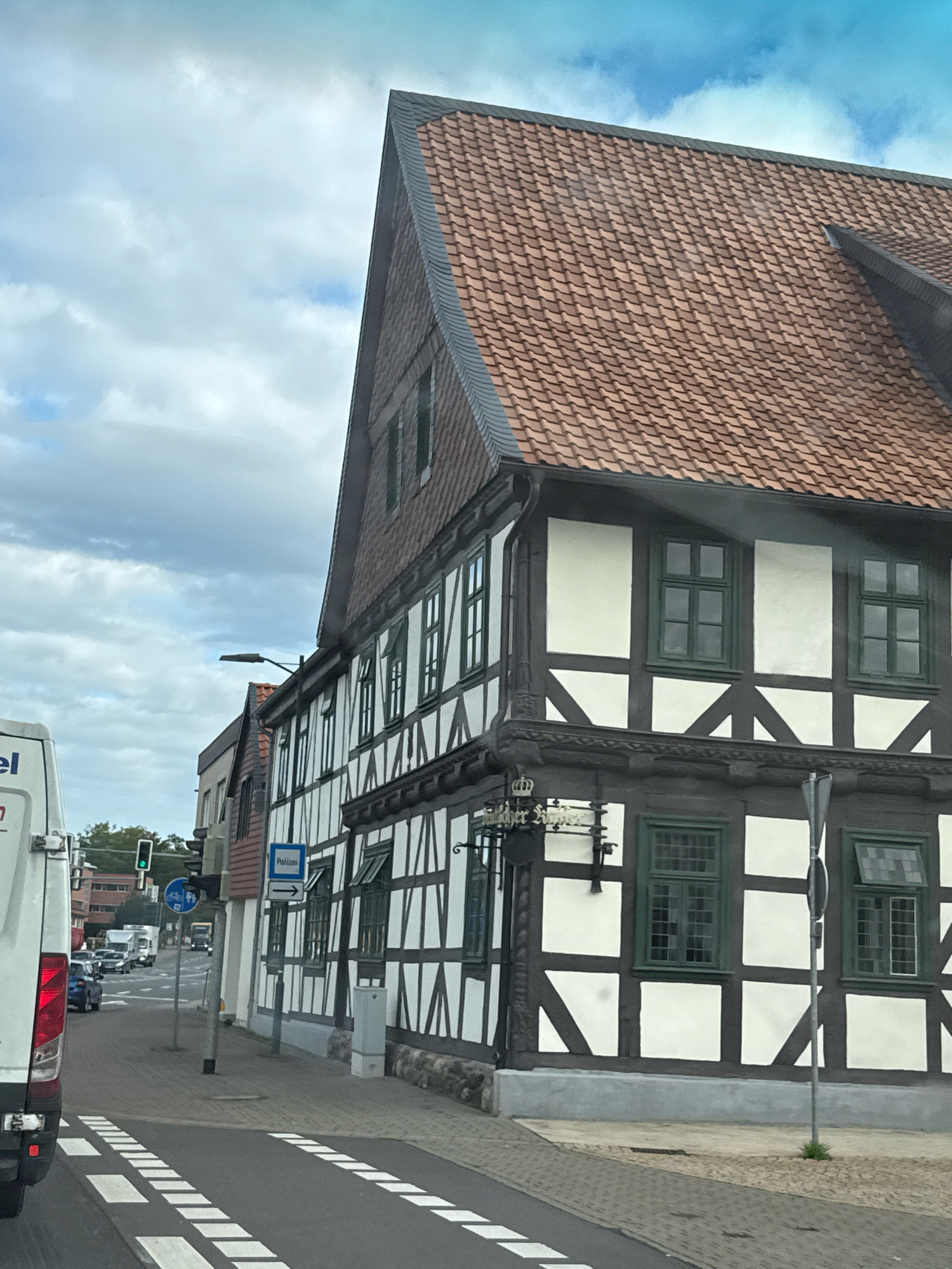 The image shows a street scene in Herzberg, Germany, featuring a traditional half-timbered building. The building has a dark brown timber frame with white infill and a red tiled roof. Several windows with dark green frames are visible on its facade. In front of the building, there is a sidewalk and a street with road markings. To the left, a white van is parked, and traffic lights and street signs are present. Cars are seen on the road in the background, and the sky is partly cloudy.