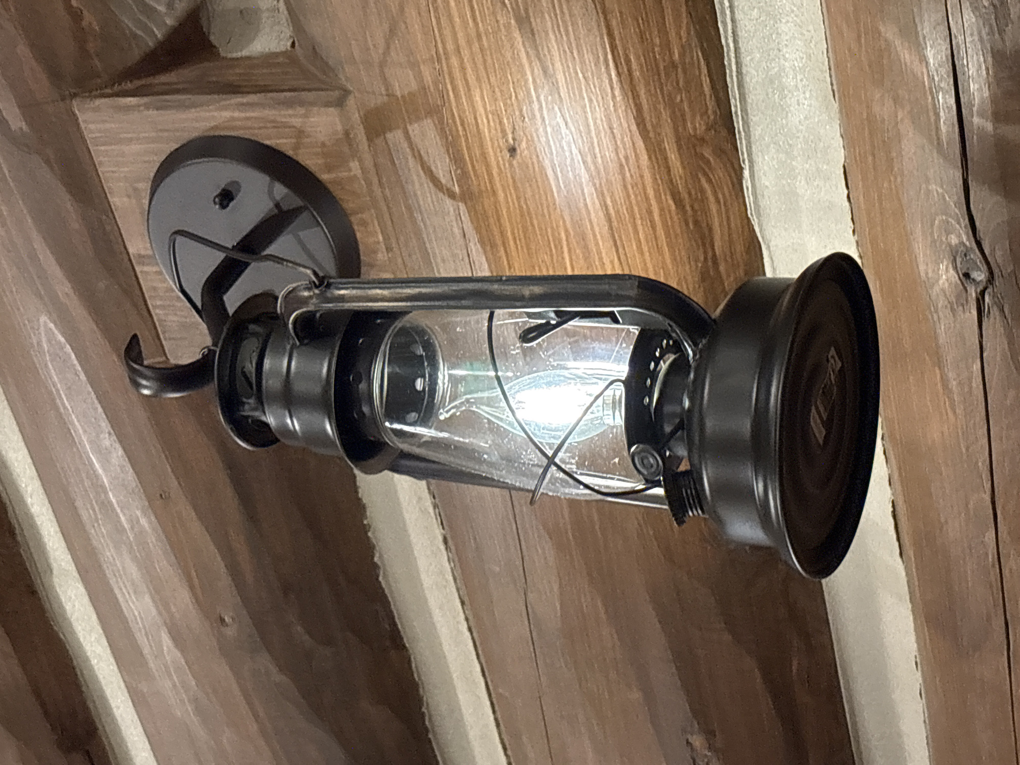 A black, kerosene-style lantern with a glass globe is mounted on a rustic wooden wall. The lantern's light bulb is illuminated. The wall is constructed from horizontally-placed, rough-hewn logs. The scene is indoors.