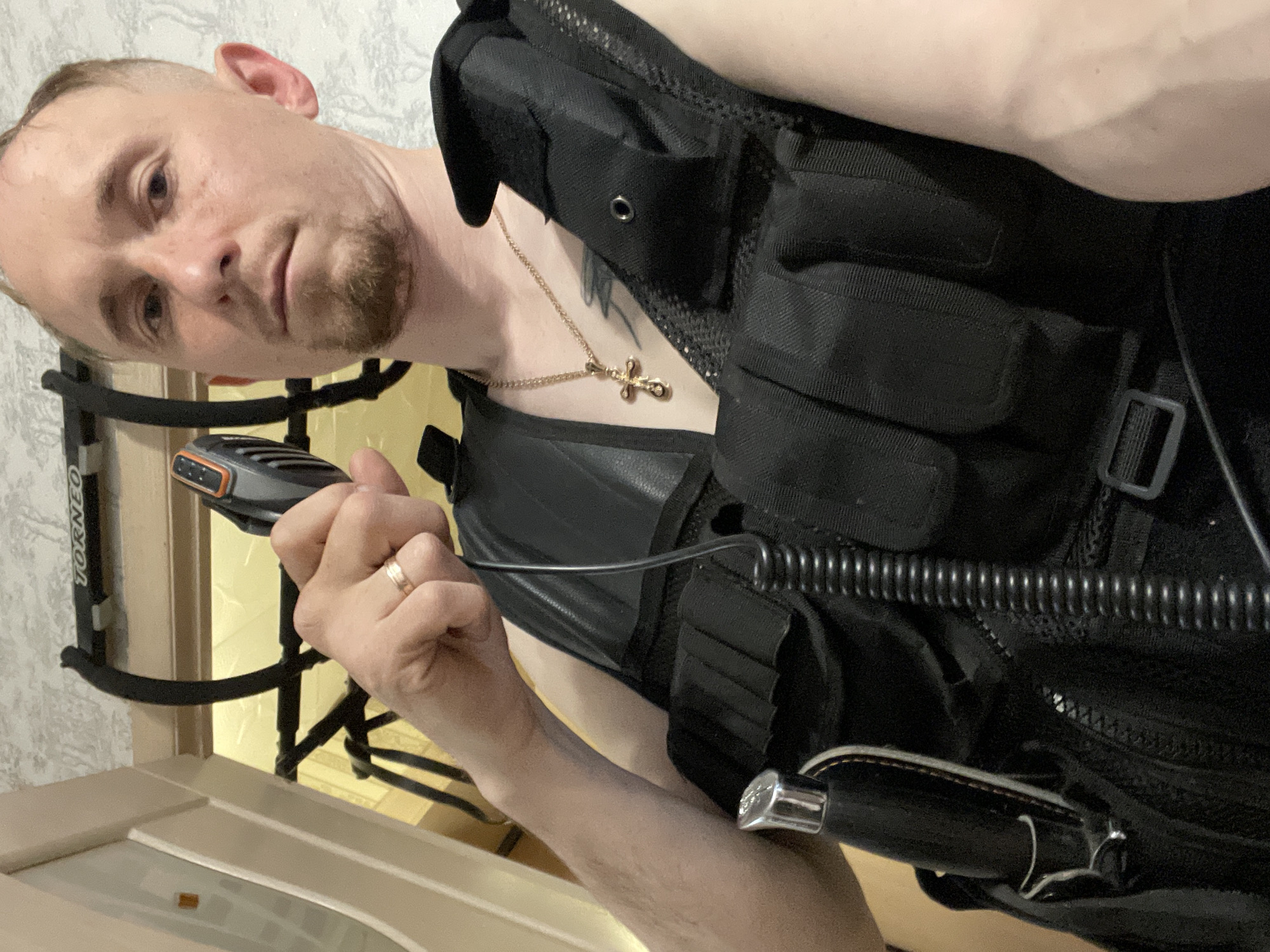 A man is shown indoors, holding a handheld microphone device with an orange accent. He is wearing what appears to be a black tactical vest with multiple pouches and a black leather vest underneath. A gold chain with a cross pendant is visible around his neck. He has short, light-colored hair and a short beard. A gold ring is visible on his ring finger. The background shows a light-colored wall with wallpaper. A piece of exercise equipment is partially visible. Part of a light-colored doorframe is also visible in the foreground. There is no information on location in the city of Sigayevo, Russia.