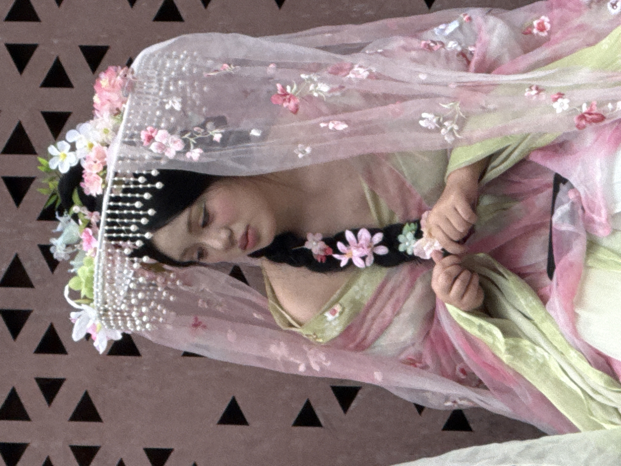 The image shows a young woman dressed in a traditional Chinese garment adorned with flowers and pearls. She is wearing a veil and her hair is braided with flowers. The background appears to be a textured wall with a geometric pattern. The woman is looking down and to the side. The overall impression is one of a ceremonial or celebratory occasion. The image does not contain enough information to determine the specific location in Yuzhong District, China.