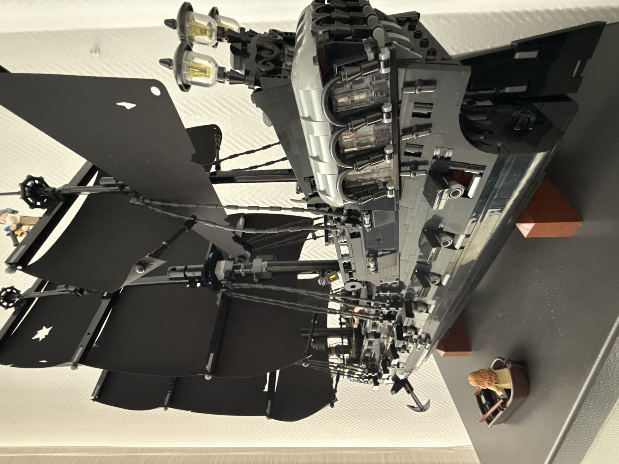 A LEGO replica of the Black Pearl pirate ship is displayed indoors. The ship is mostly black and grey, with black sails. A small dinghy with a LEGO figure is attached to the side. The ship is on display on a dark surface, with a white patterned wall in the background. The location is not specified.
