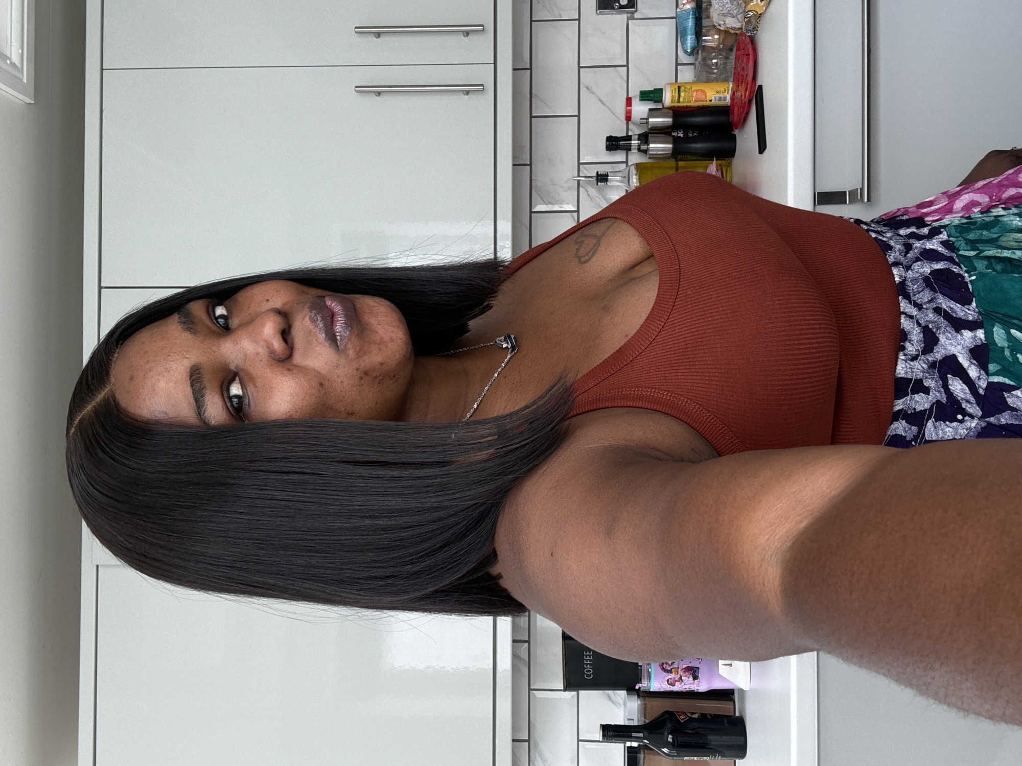 A medium shot captures a Black woman posing for a selfie in a modern kitchen. She has dark, shoulder-length hair, a heart tattoo on her upper chest, and is wearing a rust-colored, ribbed tank top and a colorful, patterned skirt. Her lips are pursed, and she is looking towards the camera. The kitchen features white subway tile backsplash, grey cabinets with silver handles, and a white countertop. On the counter are various items including a bottle of Baileys, a pink tumbler, a black coffee container, spice grinders, and bottles of olive oil and condiments. The lighting suggests an indoor setting, possibly daytime. The image does not contain any specific visual cues that would allow for its location to be identified as Darlington, United Kingdom.