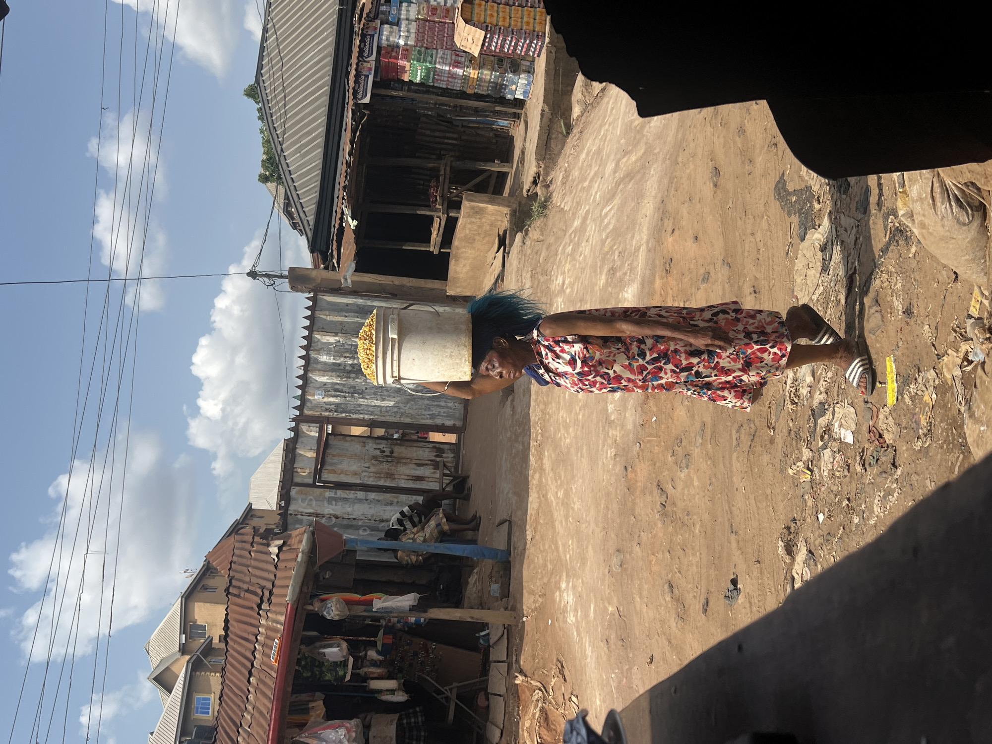 An elderly African woman carries a bucket of corn on her head down a dirt road in Onitsha, Nigeria. She wears a floral print dress and Adidas sandals. The road is lined with corrugated metal shacks selling goods, with trash visible on the ground. Overhead, a network of power lines crisscrosses the sky.