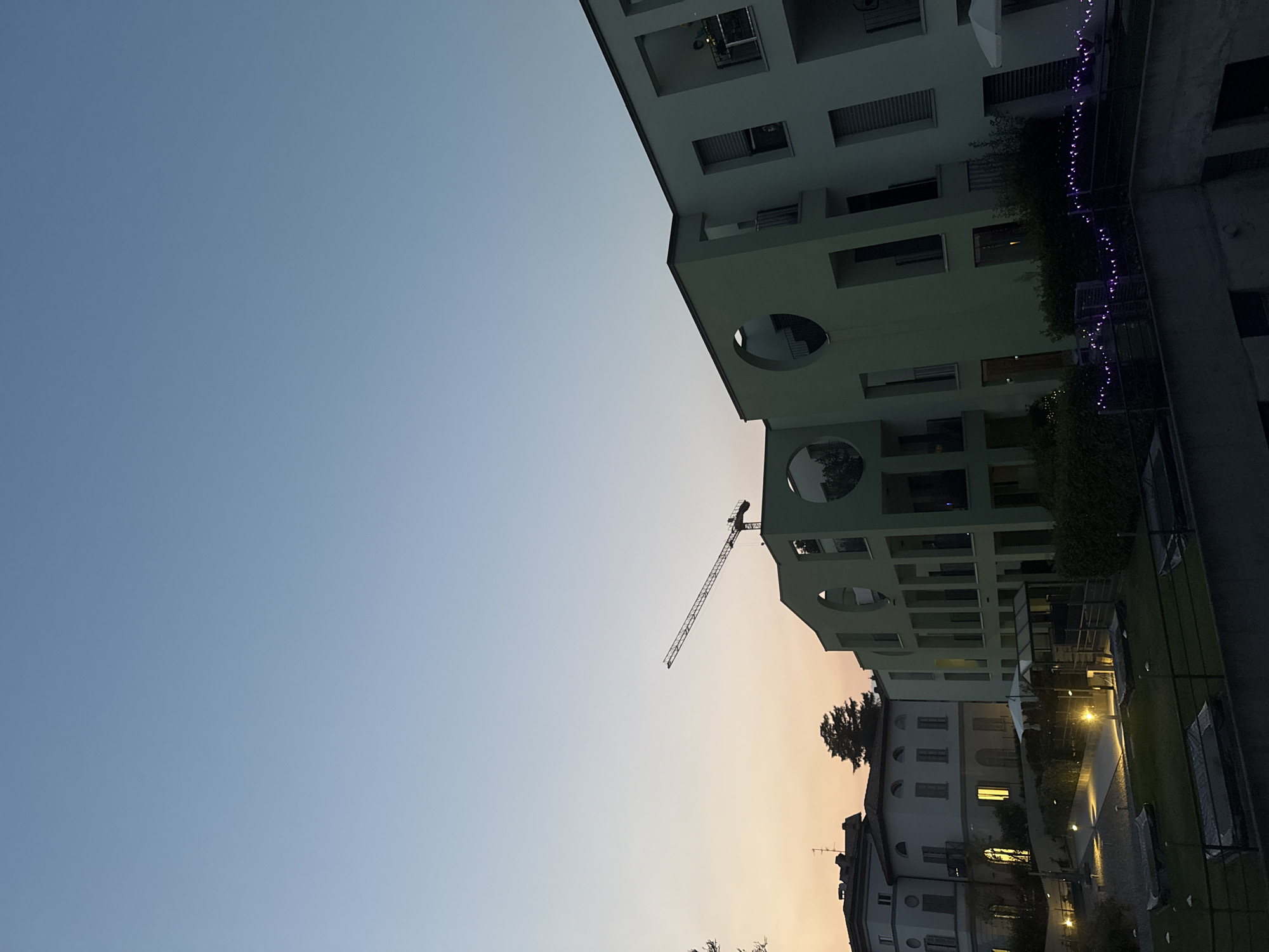In Balerna, Switzerland, at dusk, a construction crane extends over a light green, modern multi-story building. The building features circular and rectangular window openings and balconies. To the left, a more traditional, white multi-story building with arched windows is visible. The scene includes an outdoor area with illuminated pathways, vegetation, and dark-covered objects on the grass. Purple string lights are draped along a railing in the foreground. The sky is a gradient of pale blue at the top, transitioning to a soft orange and pink near the horizon.