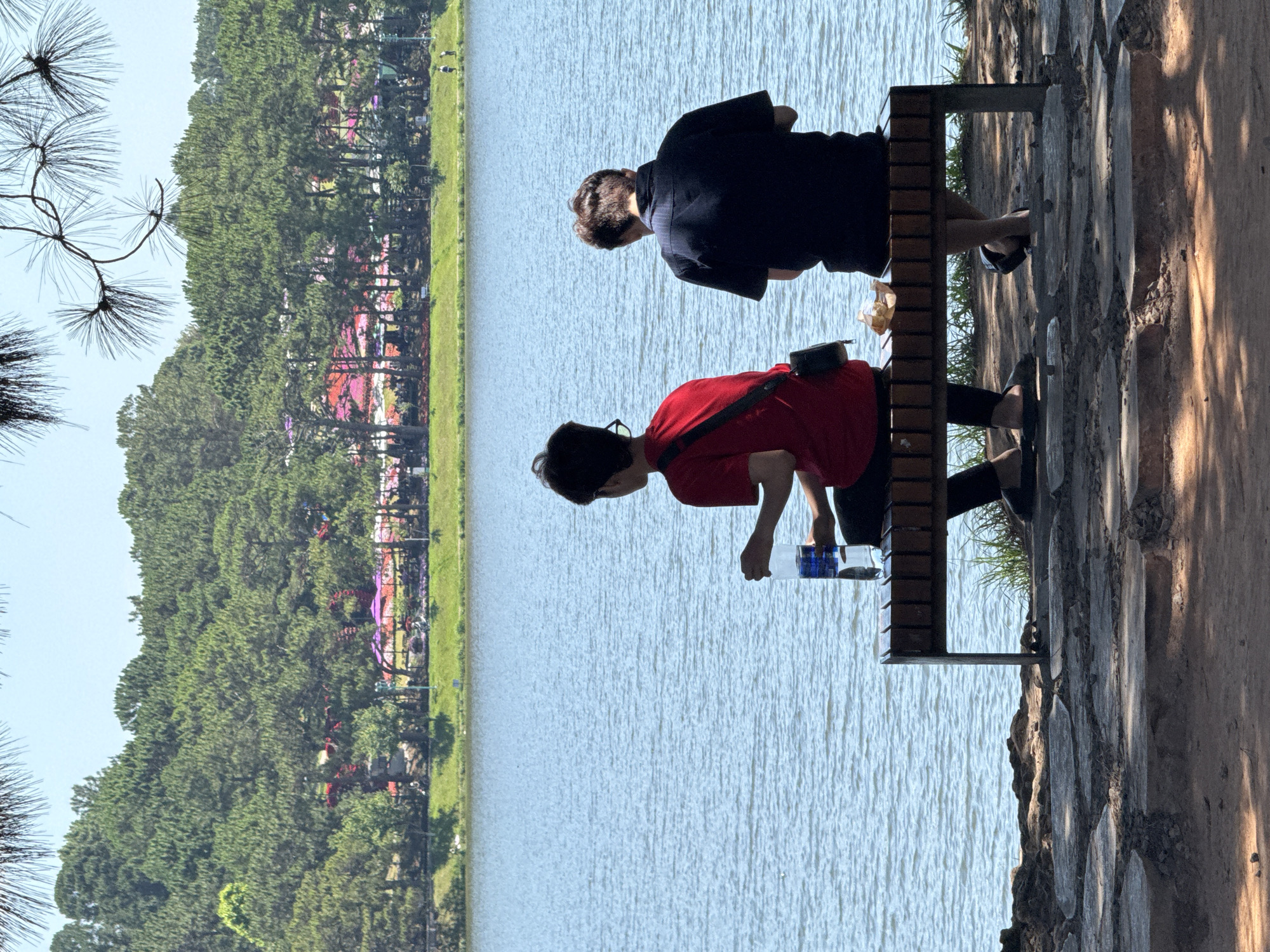 Two people are sitting on a bench looking out over a body of water. The person on the left is wearing a red shirt and has a bag slung across their chest. The person on the right is wearing a dark-colored shirt. A water bottle sits on the bench between them. The background shows a line of trees and greenery on the far shore of the water. Based on the environment and architecture, this scene is likely taken in Da Lat, Vietnam.