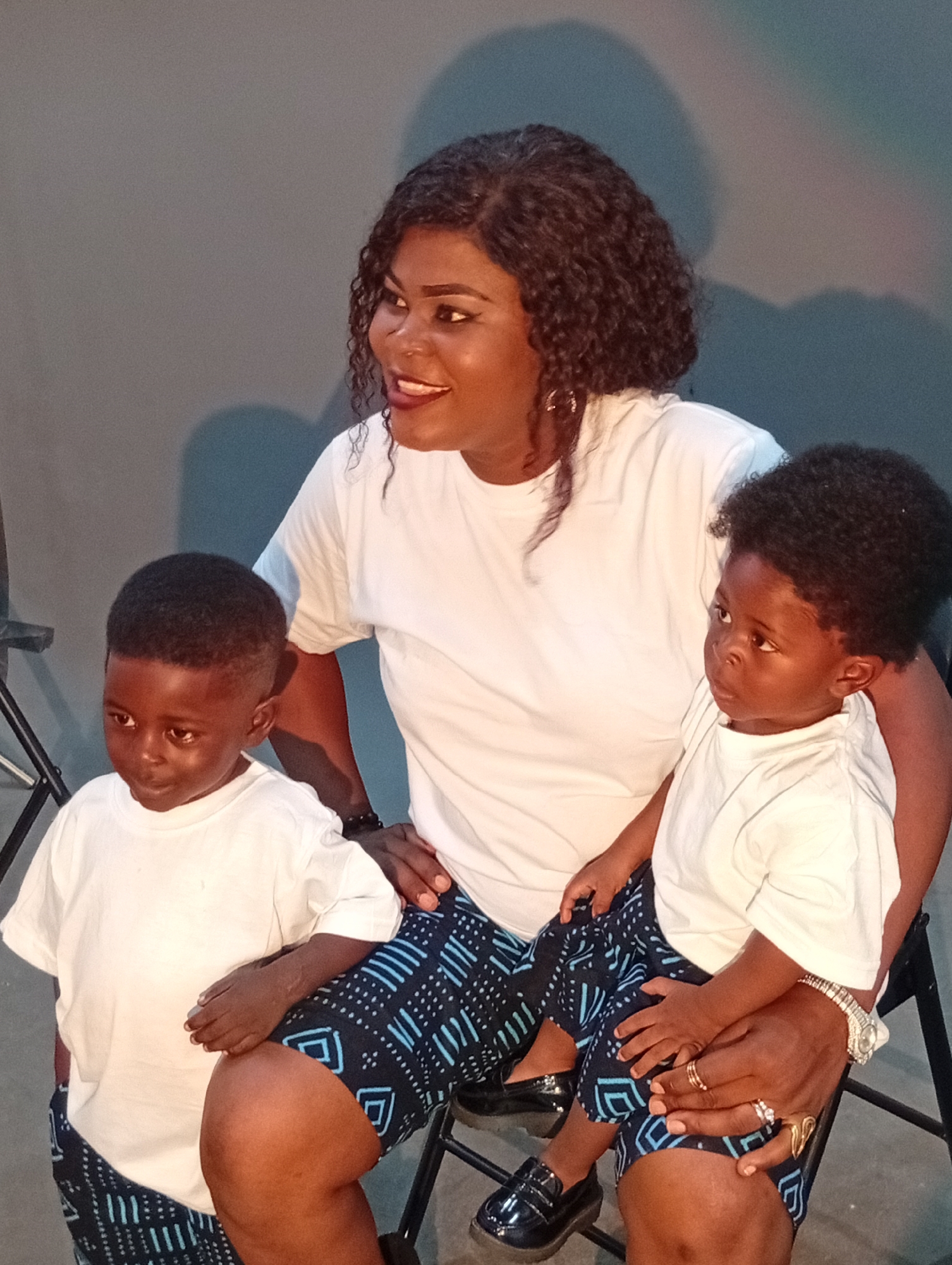A Black woman is seated, holding two young Black boys on her lap. She is wearing a white t-shirt and patterned shorts. The boys are also wearing white t-shirts and matching patterned shorts. The woman is looking to her left with her mouth open in a smile. The boy on the left is looking to his left, and the boy on the right is looking to his right. The background is a plain grey wall with a shadow of a person on it. The scene appears to be indoors.