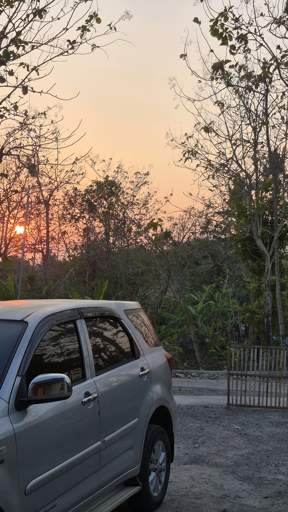 The image depicts a silver SUV parked on a gravel surface in what appears to be a rural area. The vehicle is positioned in the foreground, occupying the left half of the frame. Its side, including the rear door, rear wheel, and side mirror, is visible. Behind the car, a dense collection of trees and vegetation forms a backdrop, suggesting a natural or semi-natural environment. In the distance, a wooden fence and a gate are present. The sky above is a gradient of orange and yellow, indicating either sunrise or sunset. The sun itself is visible through the branches of trees on the left side of the image, casting a warm glow. The overall impression is of a quiet, possibly secluded location.